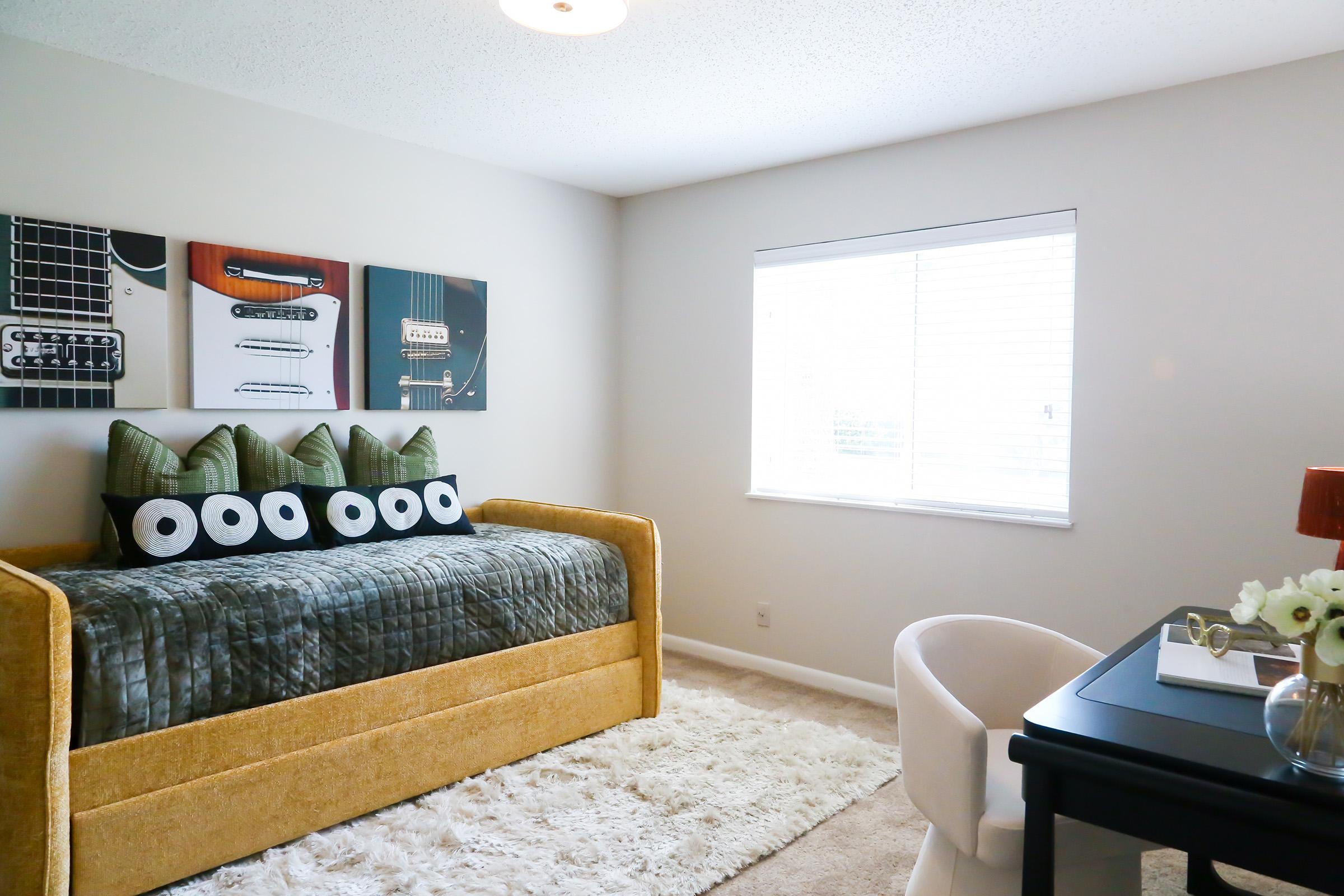 A cozy bedroom featuring a yellow daybed with green and black decorative pillows, three framed wall art pieces showcasing guitars, a white area rug, a window providing natural light, and a black desk with a white chair and a vase of flowers.