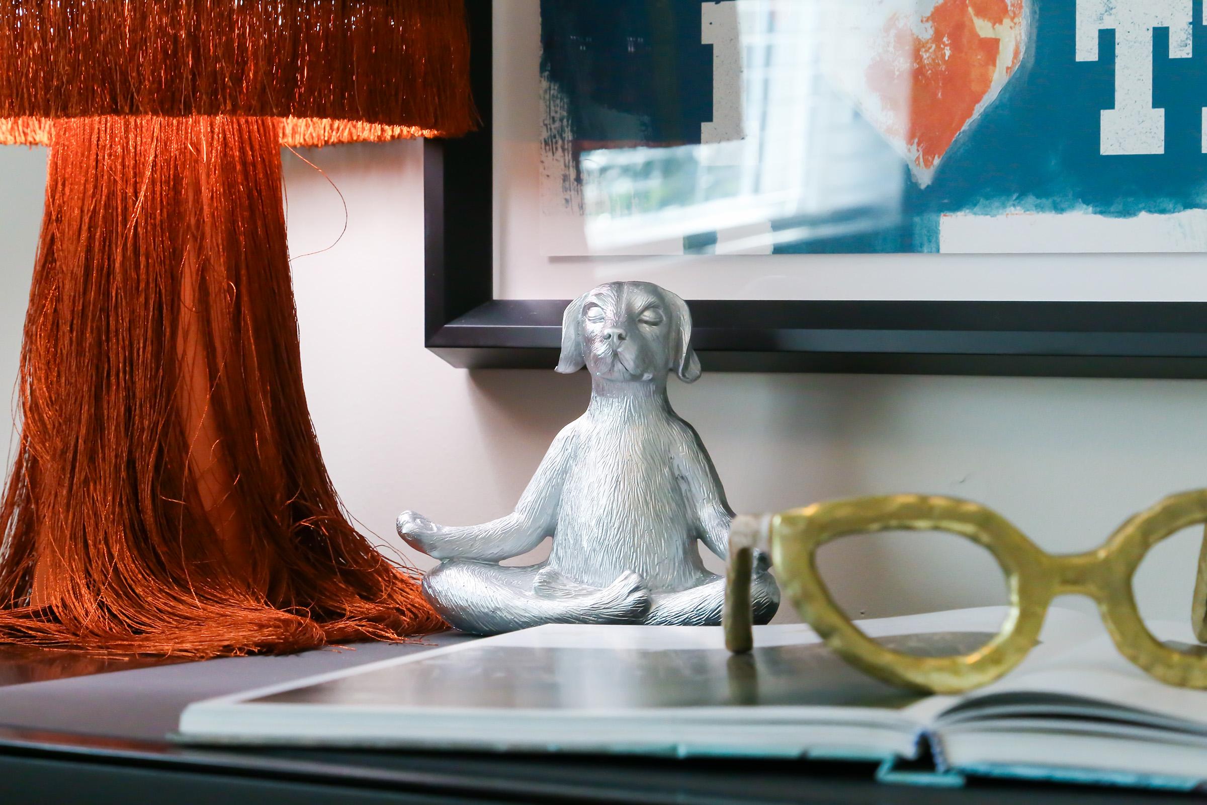 A cozy interior scene featuring a decorative silver dog figurine in a meditative pose, an orange fringe lamp, a pair of large yellow glasses, and a partially open book on a dark tabletop, with a colorful wall art piece in the background.