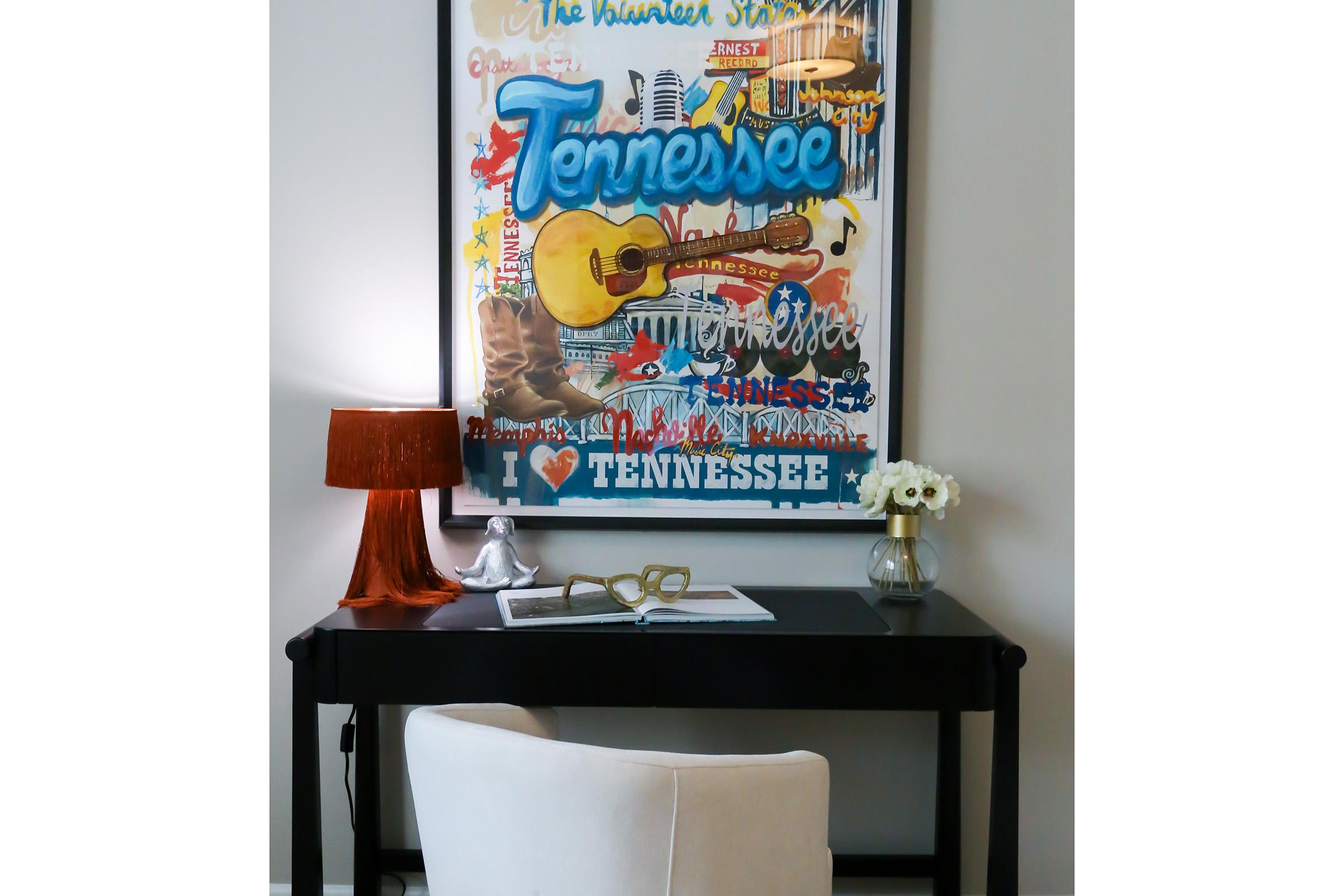 A cozy workspace featuring a black desk with a decorative lamp, a notebook, and a pair of glasses. Behind the desk, there is a colorful framed artwork titled "Tennessee," showcasing musical elements and a guitar, along with a small vase of white flowers.