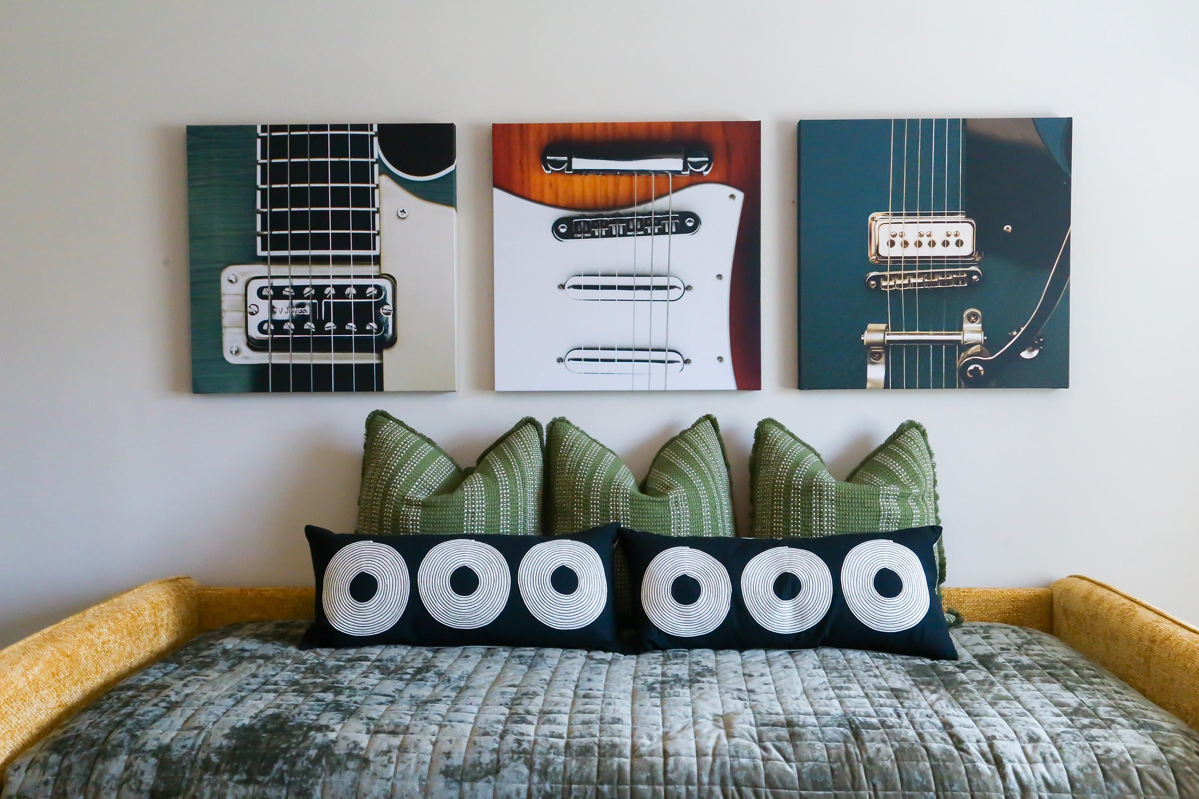 Three framed guitar photographs hang on a wall, showcasing different types of guitars. Below, a cozy couch with a quilted throw features decorative pillows with a circular pattern. The overall color scheme combines earthy tones with accents of green and yellow, creating a stylish and inviting space.
