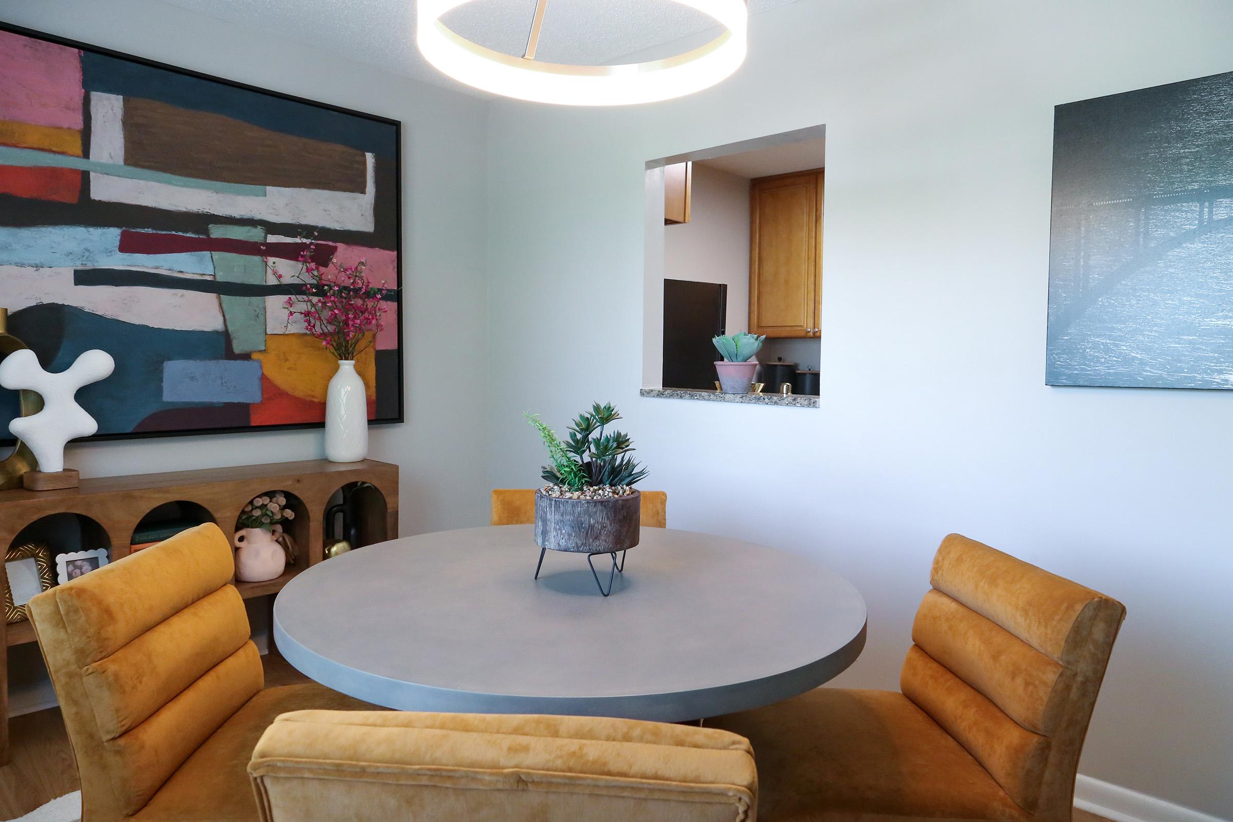 A modern dining area featuring a round gray table surrounded by four mustard-colored chairs. A decorative plant sits at the center of the table. The walls are adorned with abstract art, and a small open kitchen area can be seen in the background, showcasing wooden cabinetry and decorative plants.