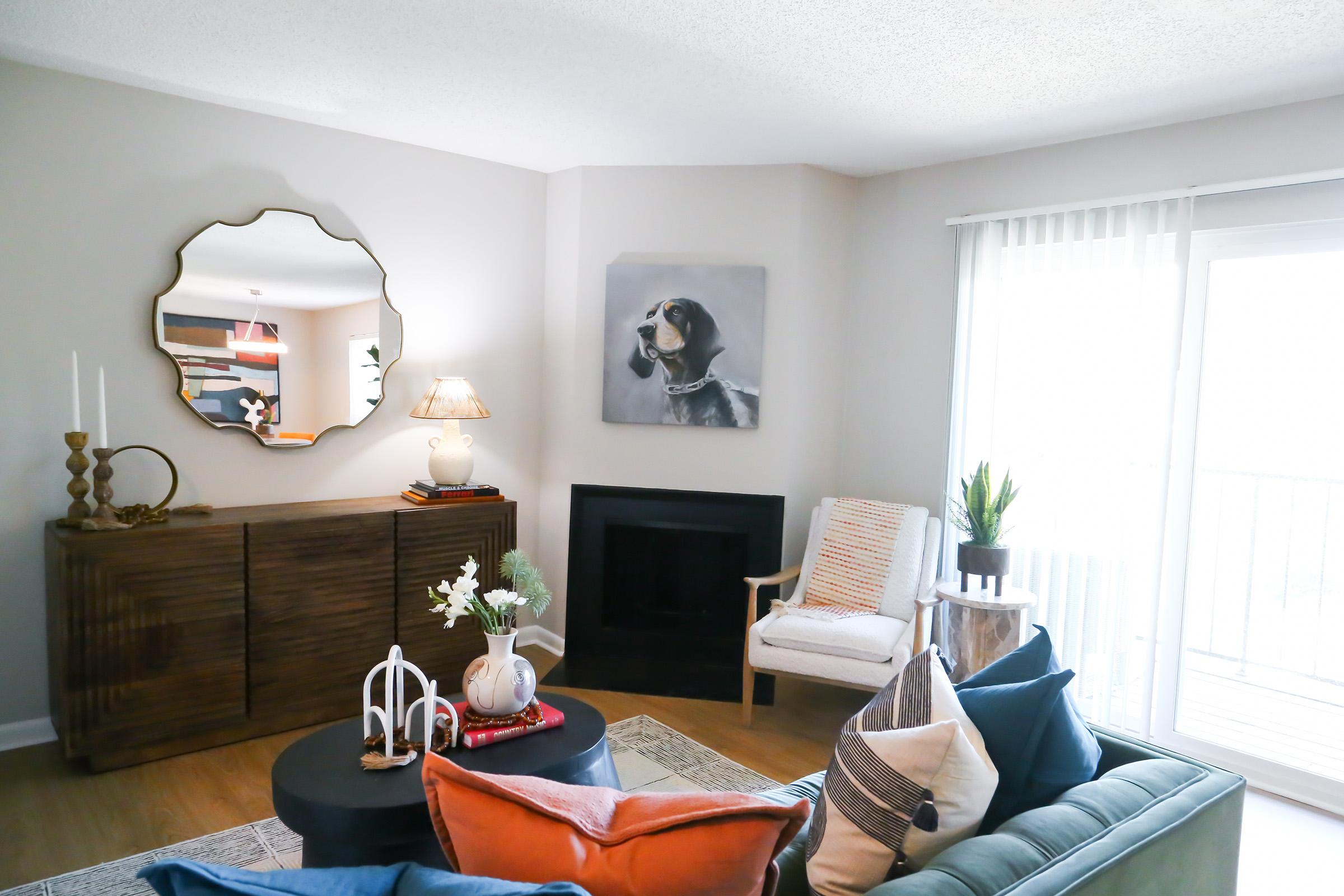 A cozy living room featuring a stylish sofa with colorful cushions, a round coffee table, and a wooden sideboard. A large mirror hangs on the wall, reflecting natural light from a sliding glass door. A painting of a dog adorns the wall, adding a personal touch to the space.