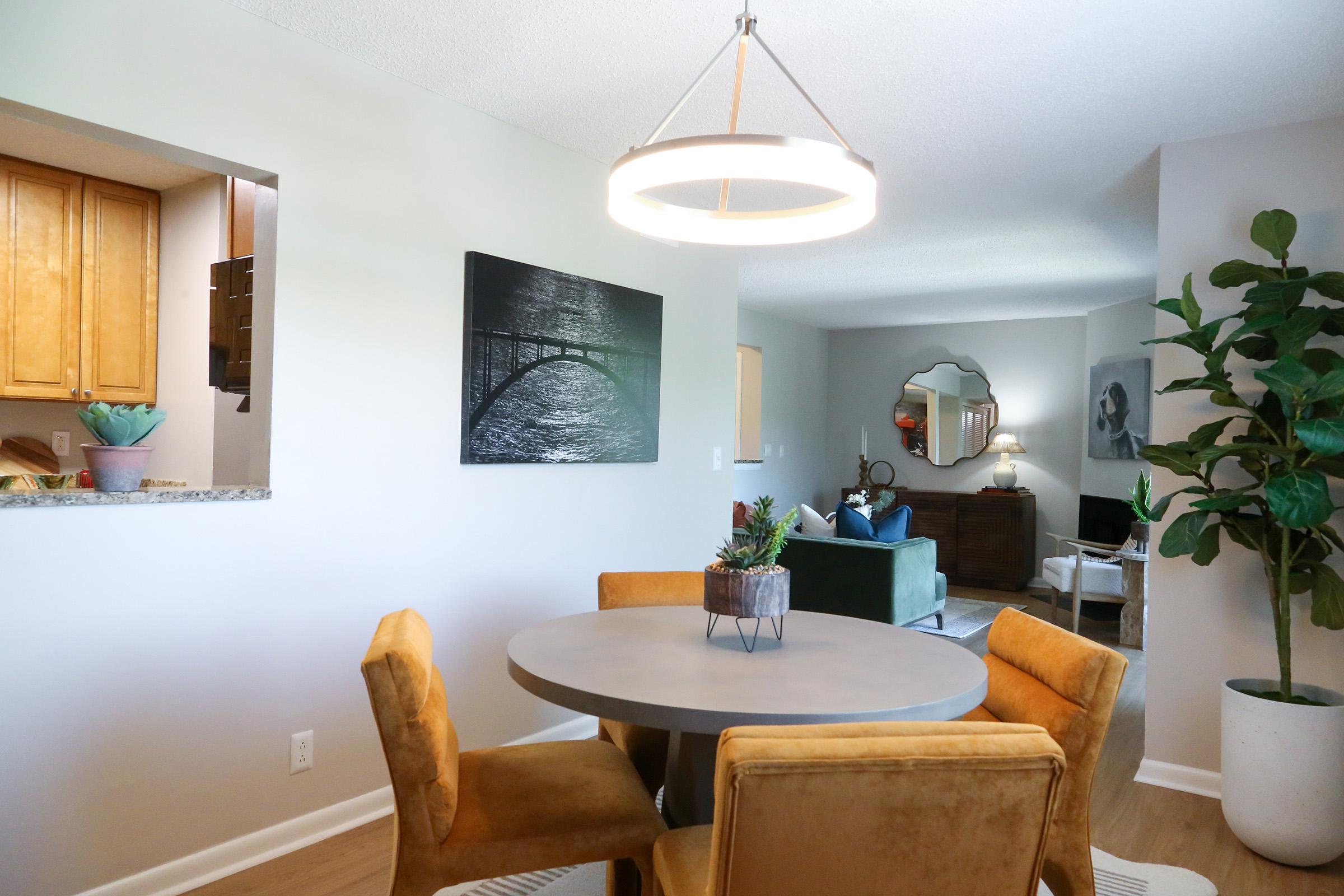 A dining area featuring a round table with four mustard-colored chairs, a small potted plant on the table, and a modern chandelier above. In the background, there's a kitchen with wooden cabinets visible through an open wall, and a living room with a green couch and decorative mirror. Bright and inviting atmosphere.