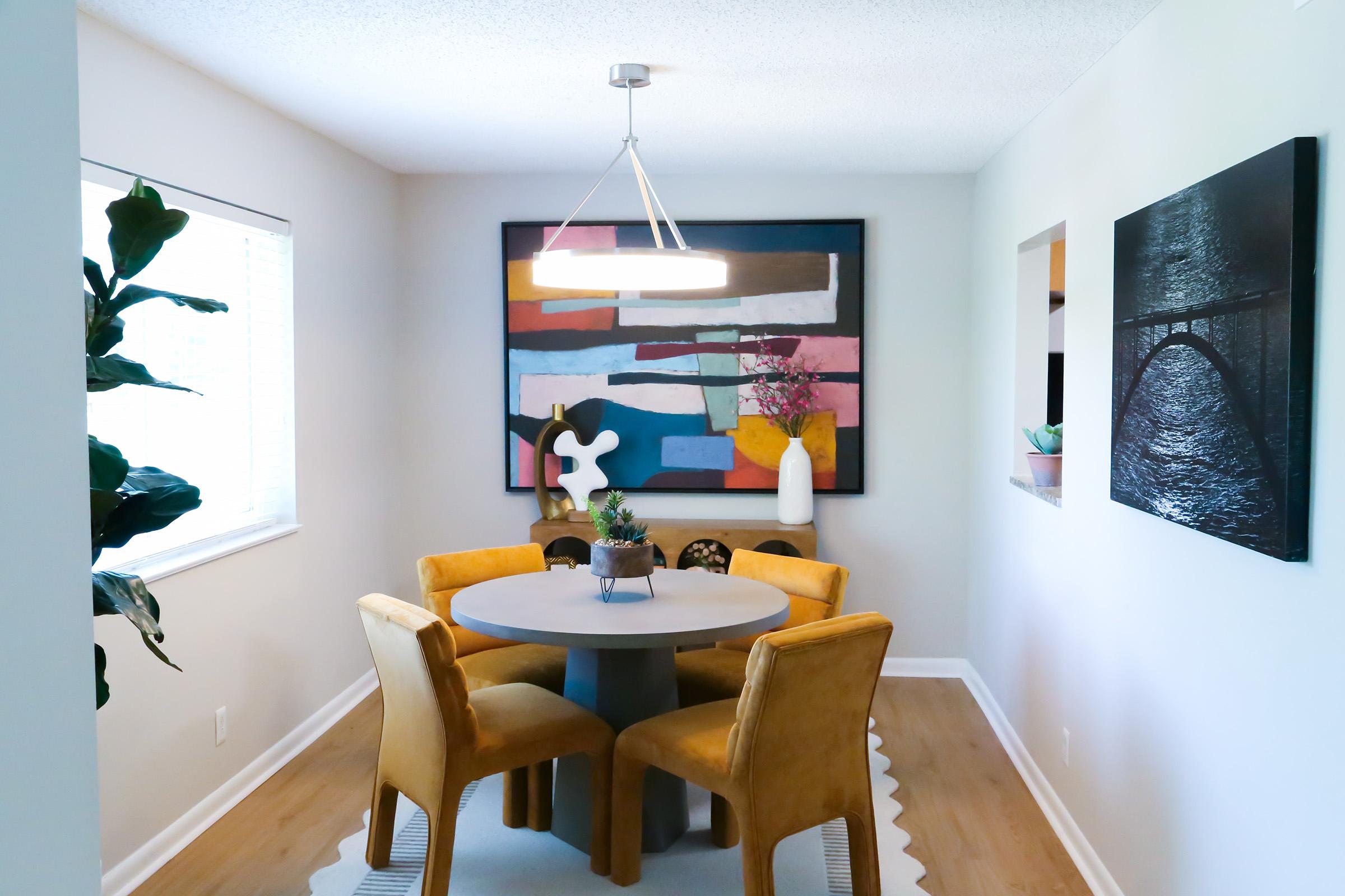A modern dining area featuring a round gray table surrounded by four yellow chairs. A colorful abstract painting hangs on the wall, complemented by decorative plants on the table and in the background. The space has soft, natural lighting and a neutral color palette, creating a warm and inviting atmosphere.
