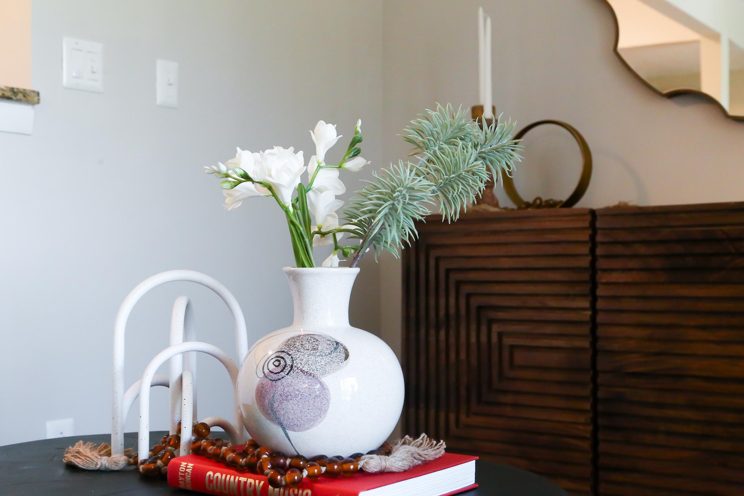 A decorative vase with white flowers and greenery sits atop a stack of books, accompanied by a wooden bead necklace and a white sculptural piece, against a neutral wall with subtle decor elements in the background.