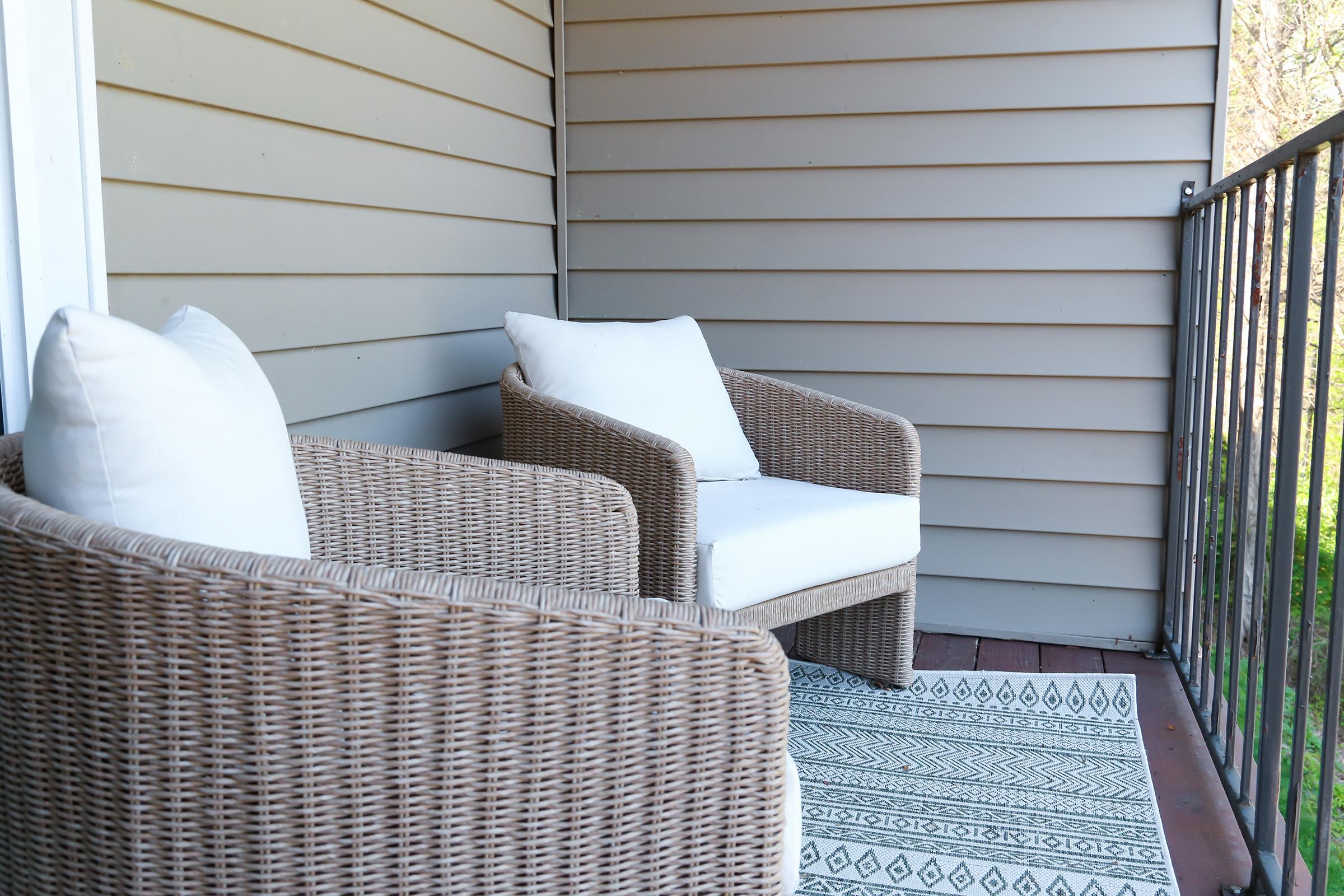 Two wicker chairs with white cushions are positioned on a wooden balcony. The chairs are placed on a patterned rug, and the background features light gray siding. The setting is bright and inviting, suggesting a cozy outdoor space for relaxation.