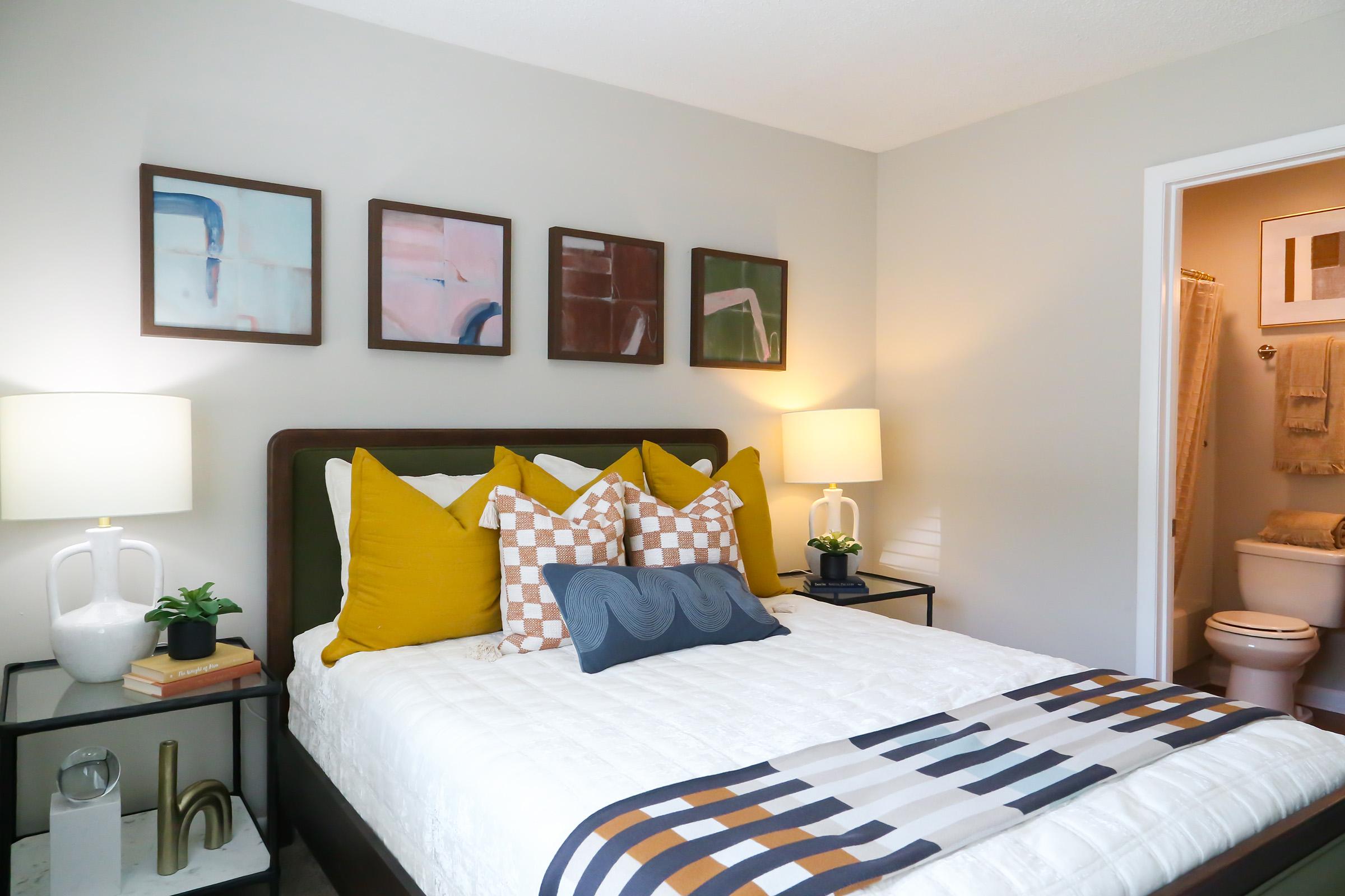 A cozy bedroom featuring a queen-sized bed with colorful throw pillows, a decorative blanket, and two bedside lamps. The walls are adorned with framed artwork, and a doorway leads to a bathroom in the background, hinting at a modern design aesthetic. A small plant adds a touch of greenery.
