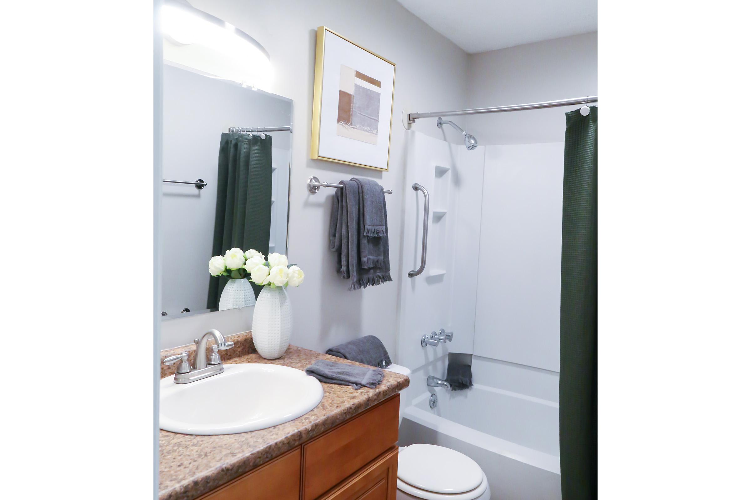 A well-lit bathroom featuring a white bathtub with a shower, a large mirror above a sink, and a wooden vanity. Decor includes a wall art piece in a frame, a vase with white flowers, and a few towels hung neatly. The color scheme combines neutral tones with dark green accents.