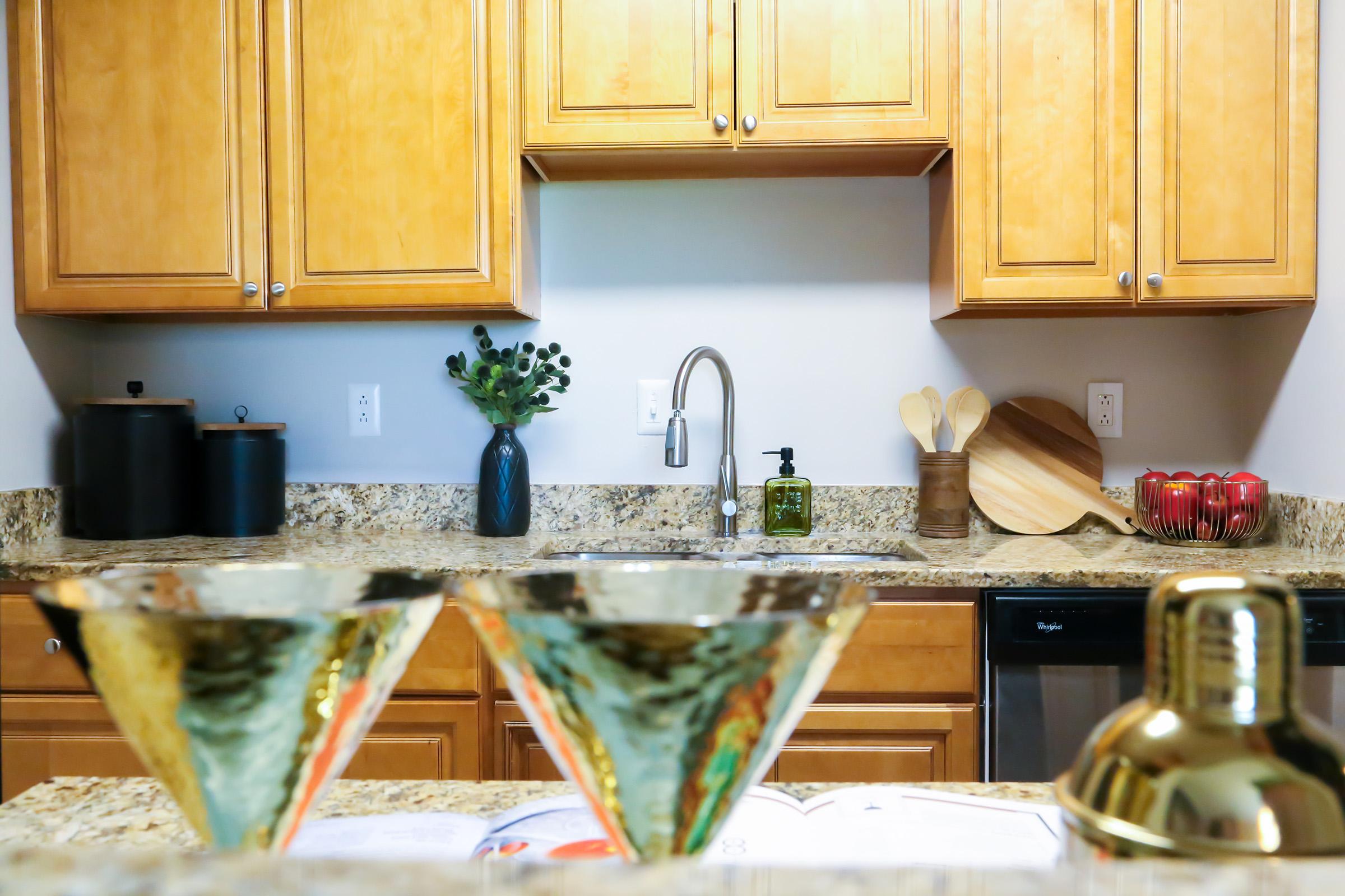 Modern kitchen with wooden cabinets, a granite countertop, and various kitchen accessories. There are two glass bowls in the foreground, a sink with a sleek faucet, a potted plant, and wooden utensils. The setting is bright and inviting, conveying a cozy atmosphere.