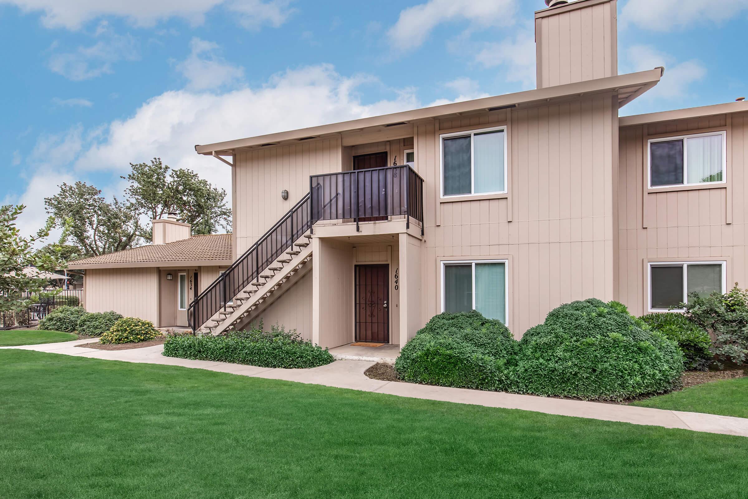 A two-story apartment building with a staircase leading to a second-floor entrance. The building features beige siding, several windows, and green shrubs in the front yard. A well-maintained lawn surrounds the pathway leading to the entrance. The sky is partly cloudy, creating a pleasant atmosphere.
