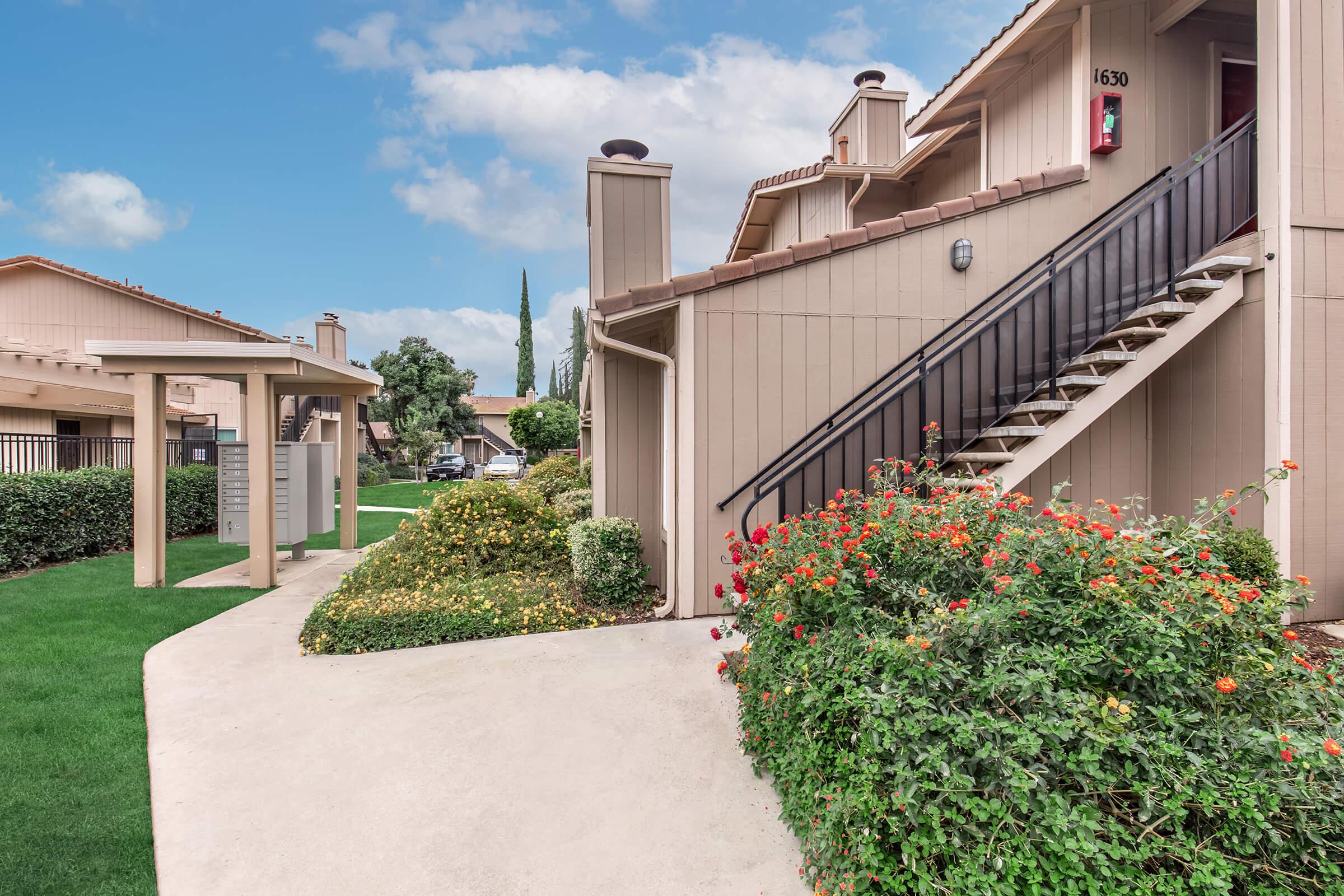 Photo of a residential building exterior featuring a staircase leading to an upper floor, with a pathway lined by vibrant flower bushes. In the background, there are trees and additional buildings visible under a partly cloudy sky. The setting appears well-maintained and inviting.