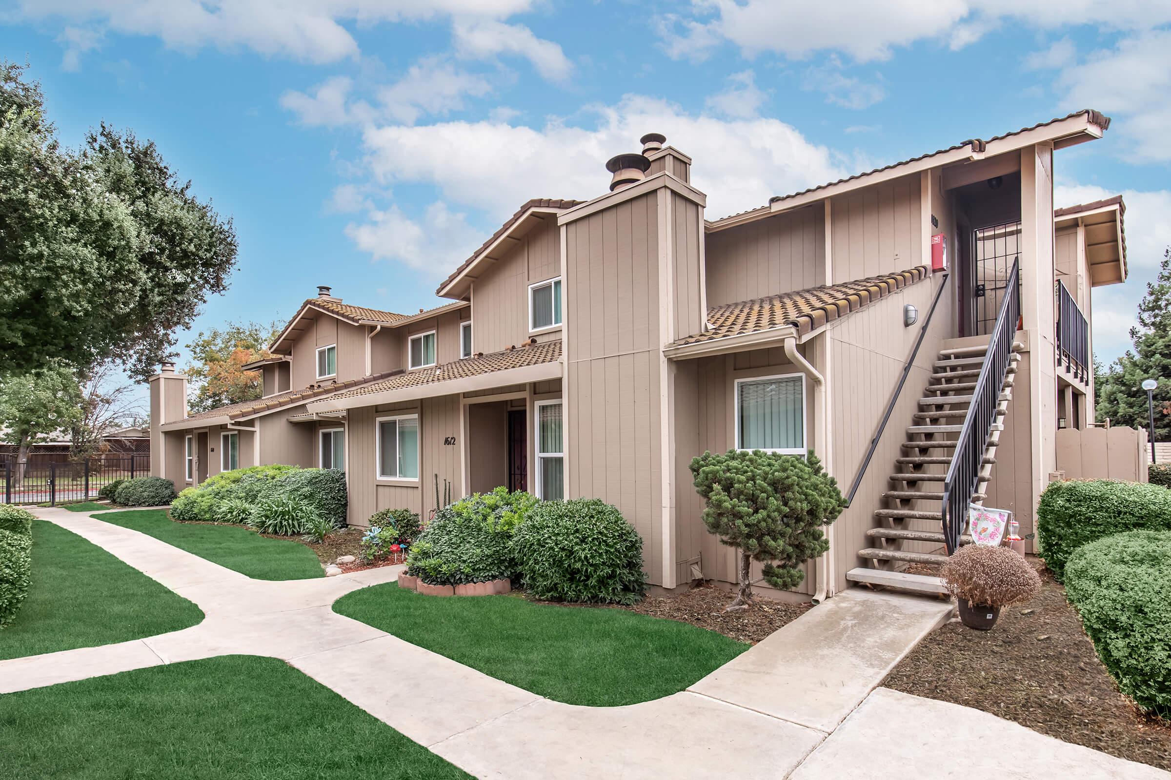 A two-story apartment building with a light brown exterior, featuring a tiled roof and multiple windows. The building is surrounded by well-maintained green lawns and small shrubs, with a concrete pathway leading to the entrance. Clear blue sky with some clouds in the background.