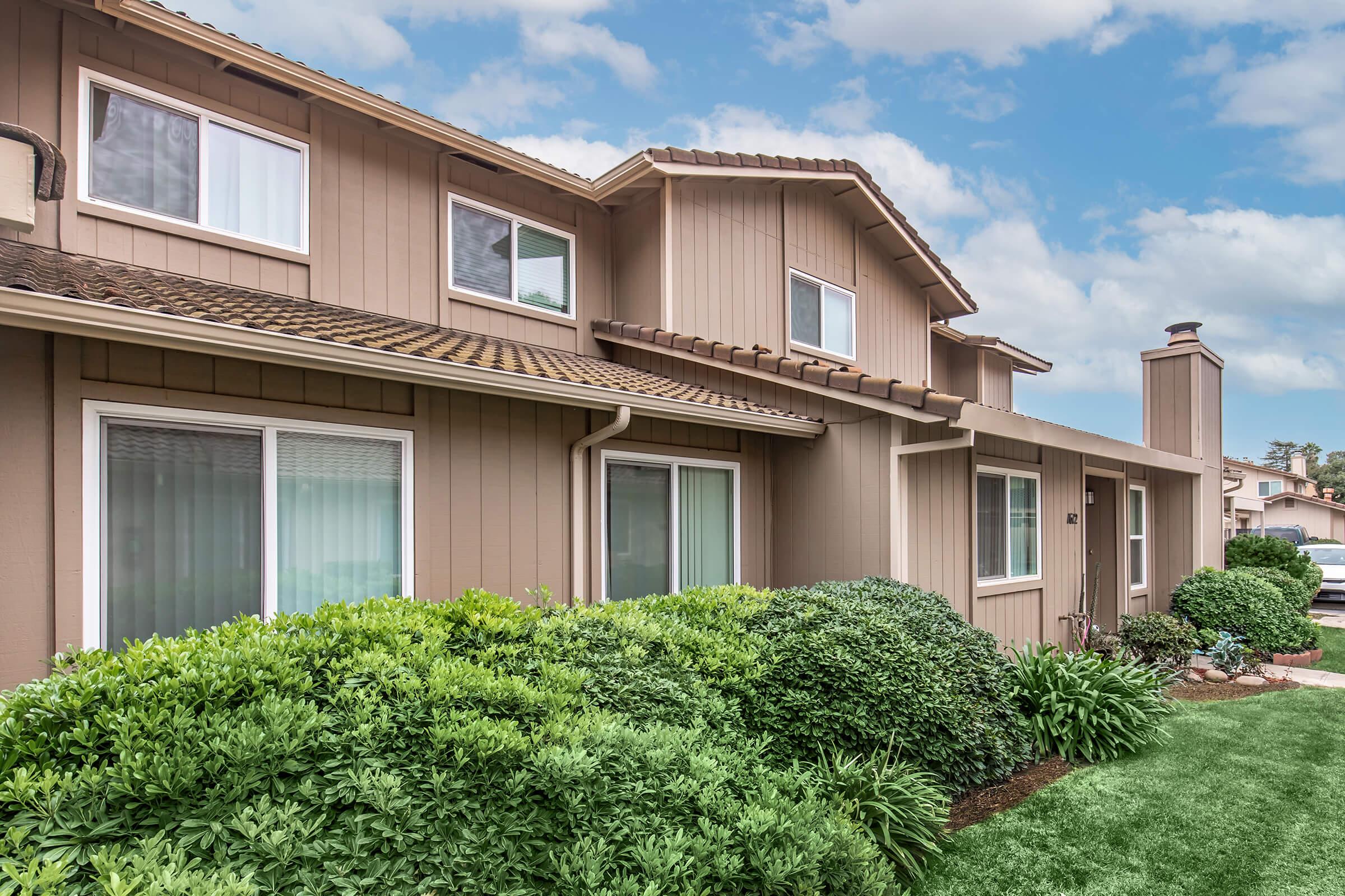 A view of a two-story house featuring a light brown exterior, multiple windows, and a well-maintained landscape with lush green shrubs in the foreground. The sky is partly cloudy, adding a calm ambiance to the suburban setting.