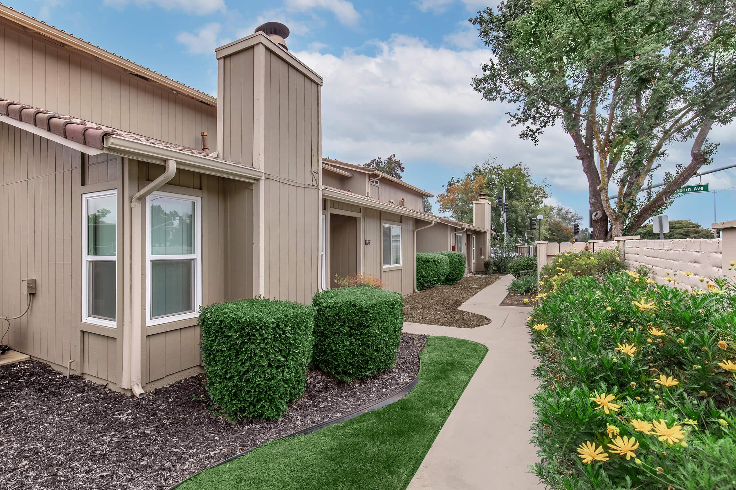 A well-maintained pathway leading through a residential area, flanked by two-story beige buildings. Neatly trimmed shrubs line the path, alongside colorful flowers and greenery. Trees and blue sky are visible in the background, contributing to a pleasant outdoor atmosphere.