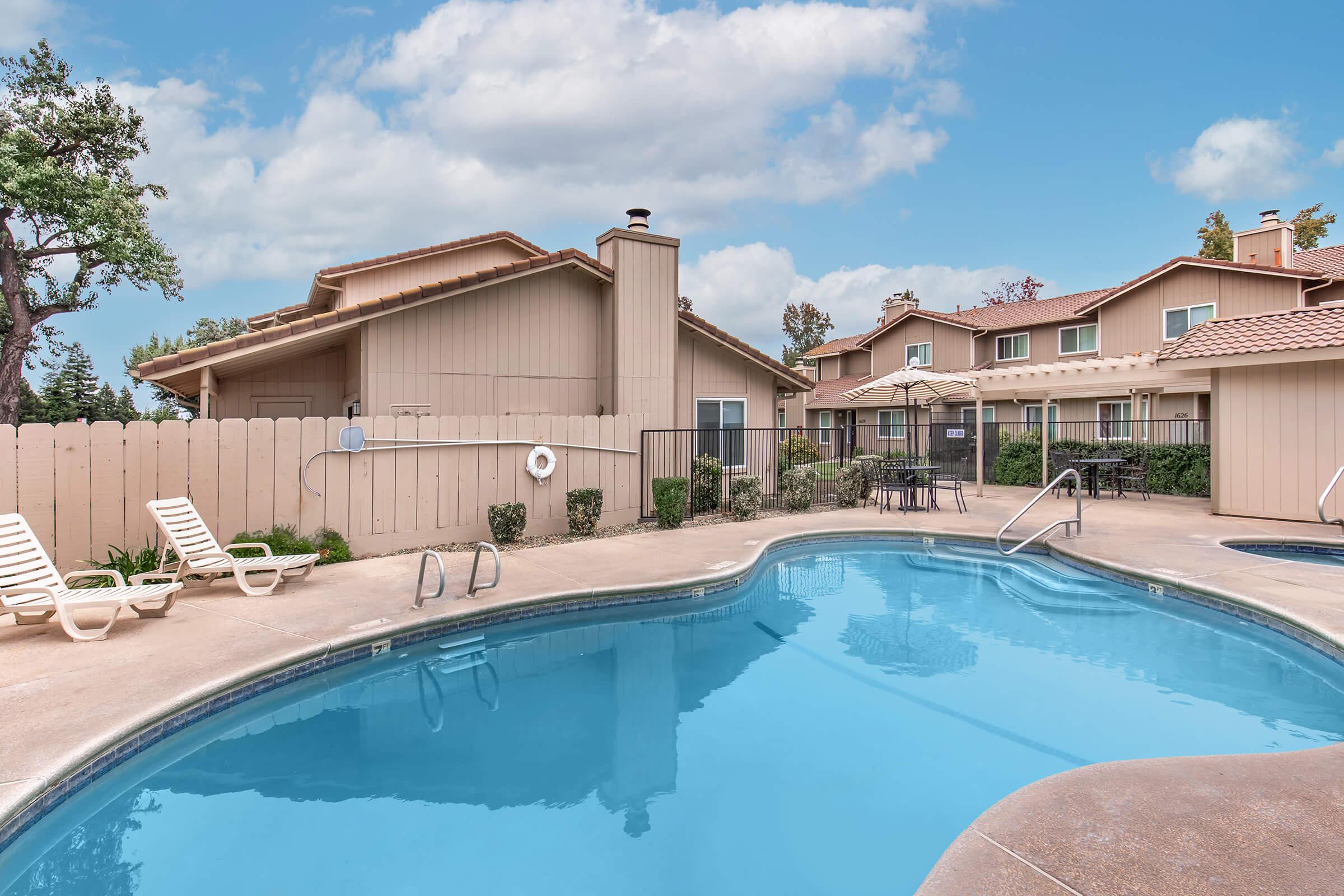 A swimming pool surrounded by lounges and tables, with buildings visible in the background. The sky is partly cloudy, creating a serene atmosphere. Lush greenery is present around the pool area, enhancing the outdoor space's appeal.