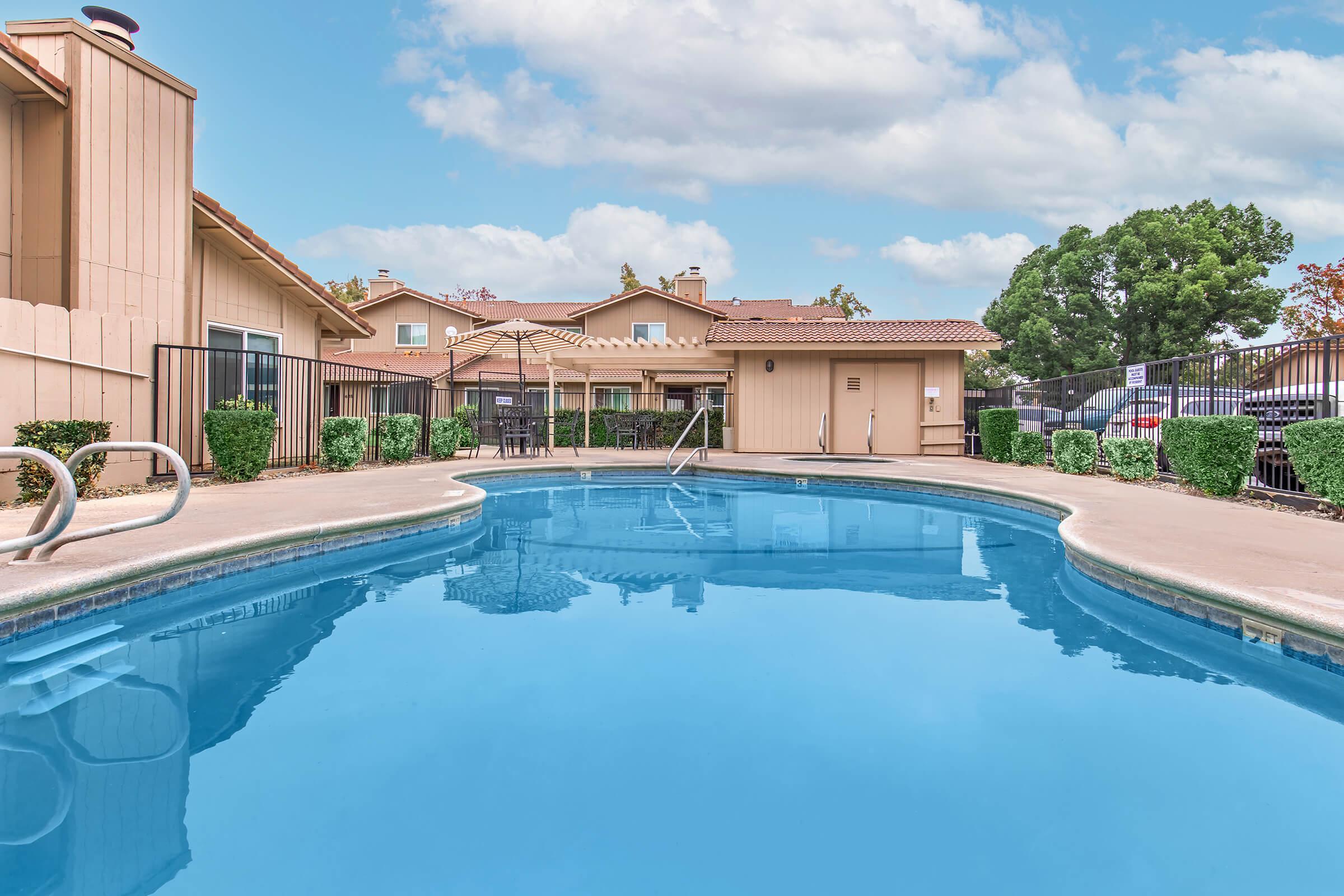 A clear blue swimming pool surrounded by a neatly landscaped area with small bushes. In the background, there are residential buildings and a pergola, with a bright sky partially covered by fluffy clouds. The pool area features a fence for safety and a set of pool steps.
