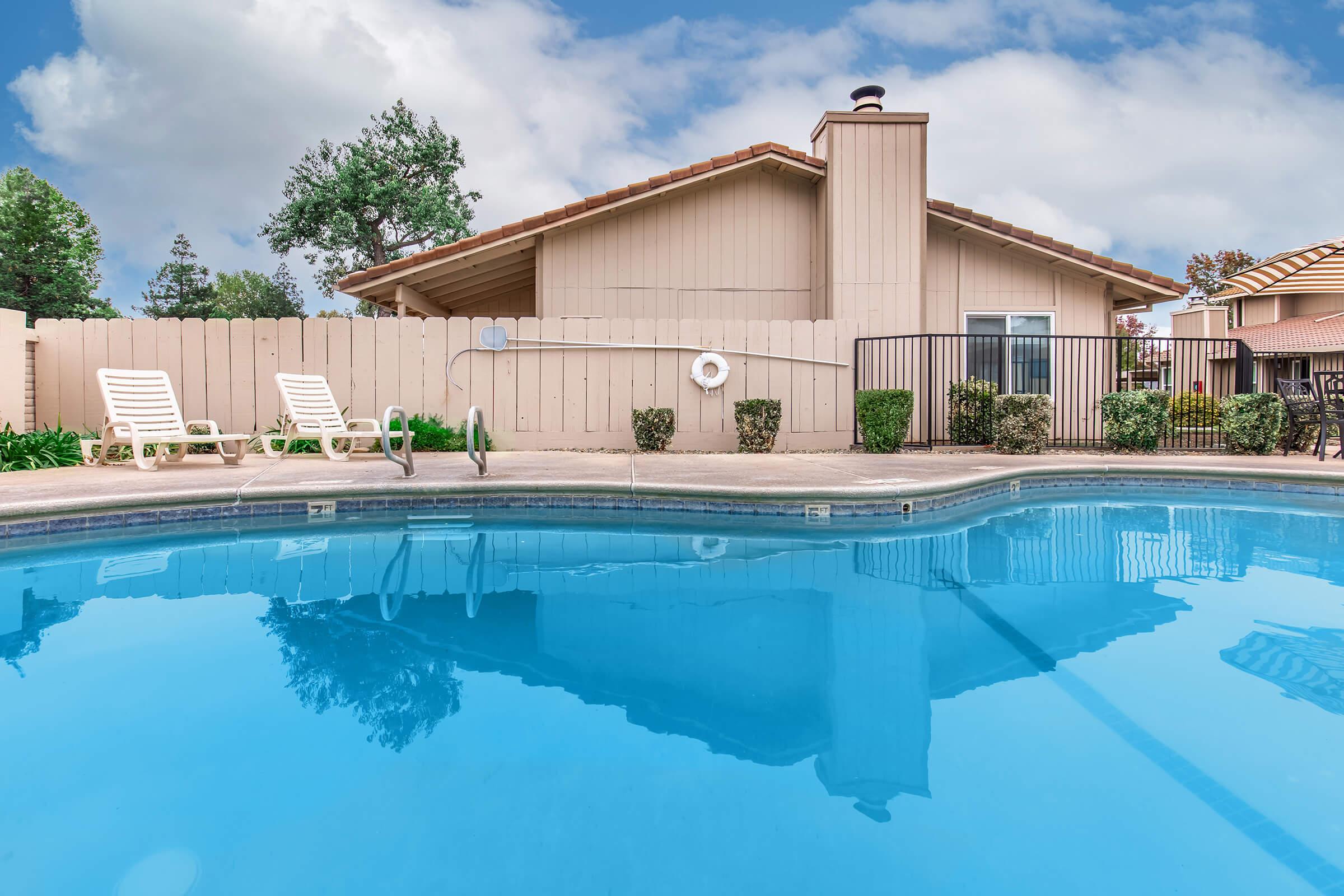 A clear blue swimming pool with reflective water, surrounded by a concrete deck. There are two white lounge chairs and a wooden fence in the background, with a beige house featuring a chimney and landscaped greenery nearby. The sky is partly cloudy, enhancing the serene atmosphere.