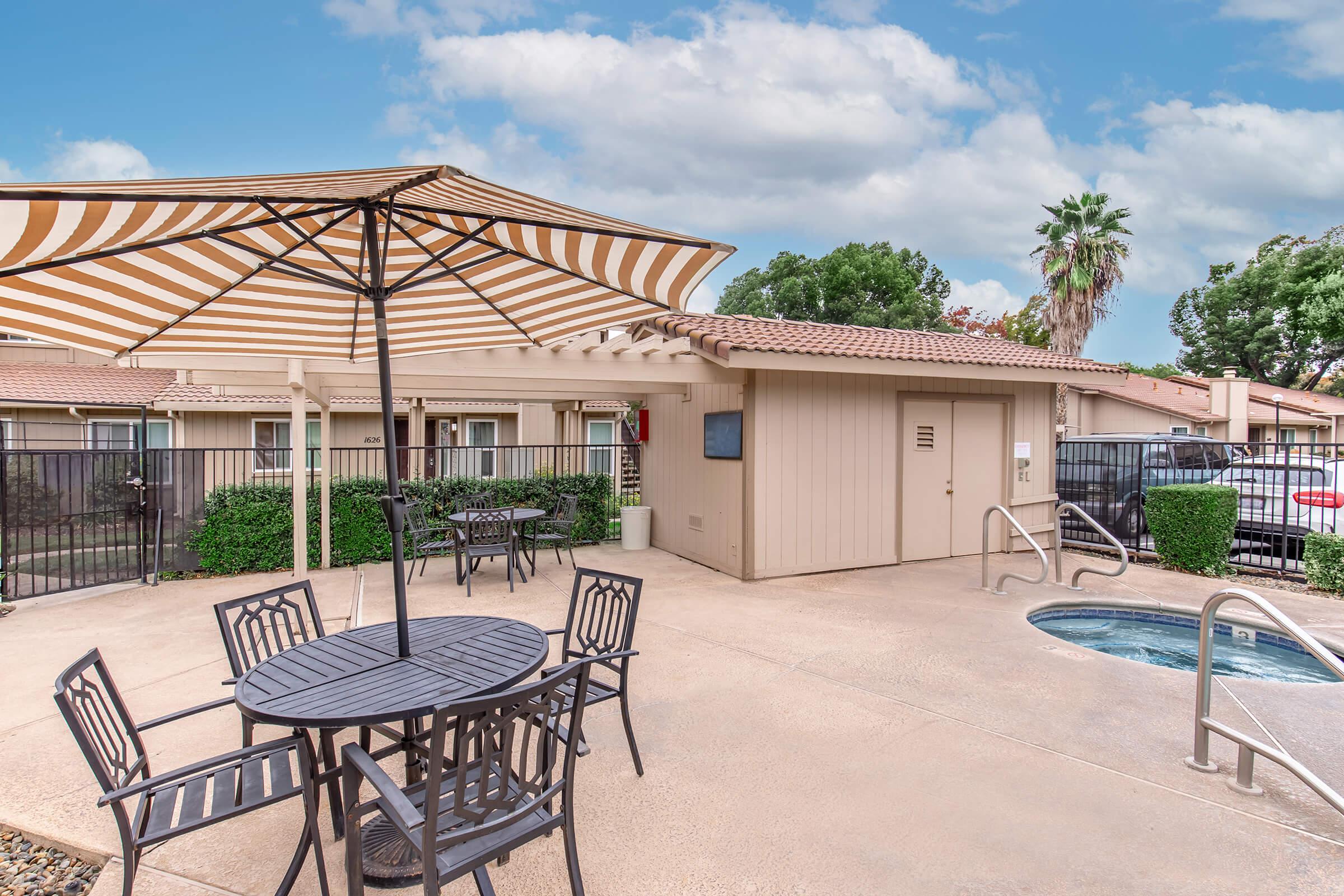 Outdoor seating area featuring a shaded table and chairs beside a hot tub. The space is surrounded by greenery, with a backdrop of residential buildings and palm trees under a partly cloudy sky.