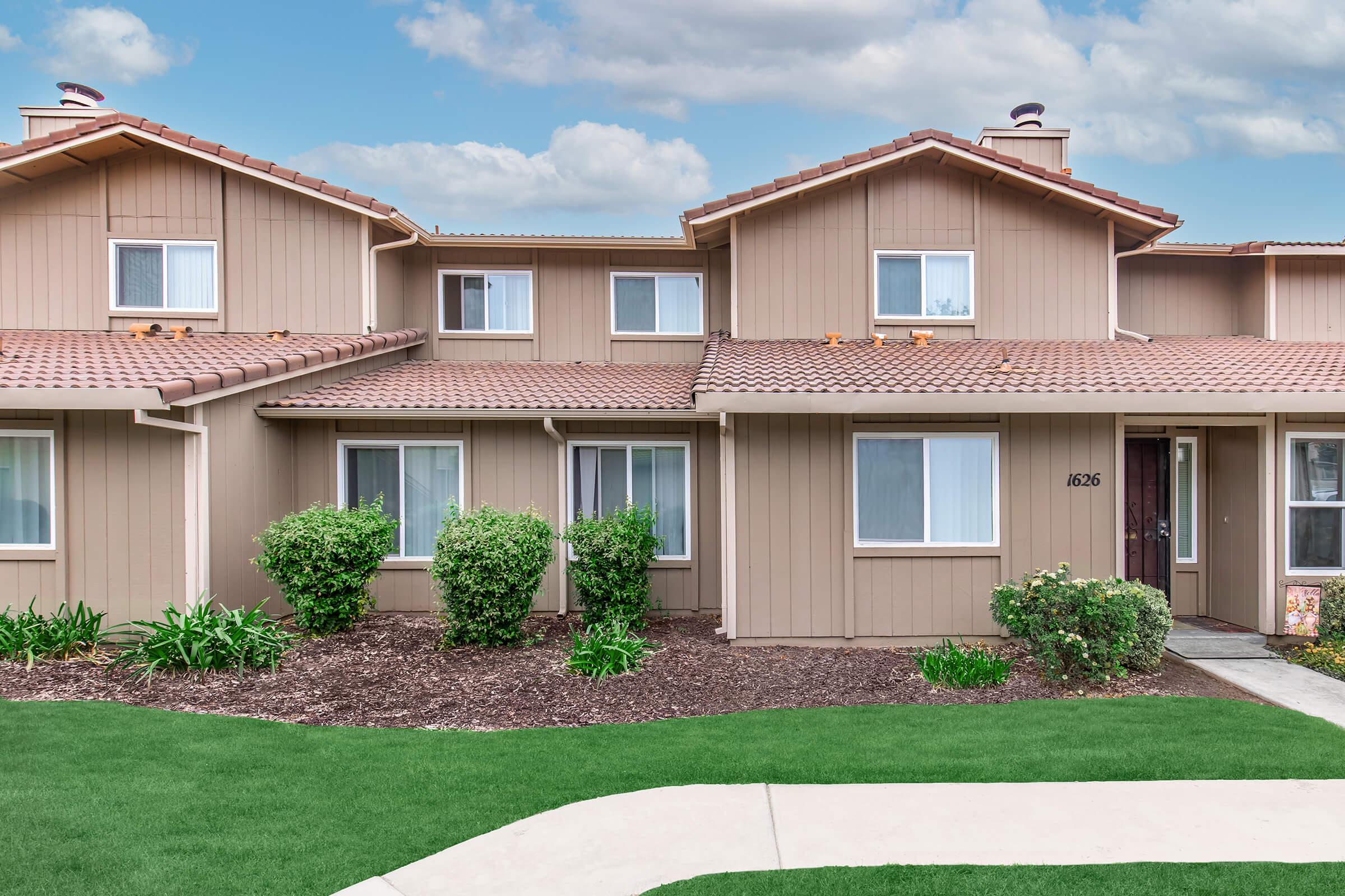 A well-maintained beige townhouse with a brown tiled roof. The front yard features manicured green grass, neatly trimmed bushes, and small plants. The structure has multiple windows and a clean pathway leading to the entrance, set against a background of partly cloudy skies.