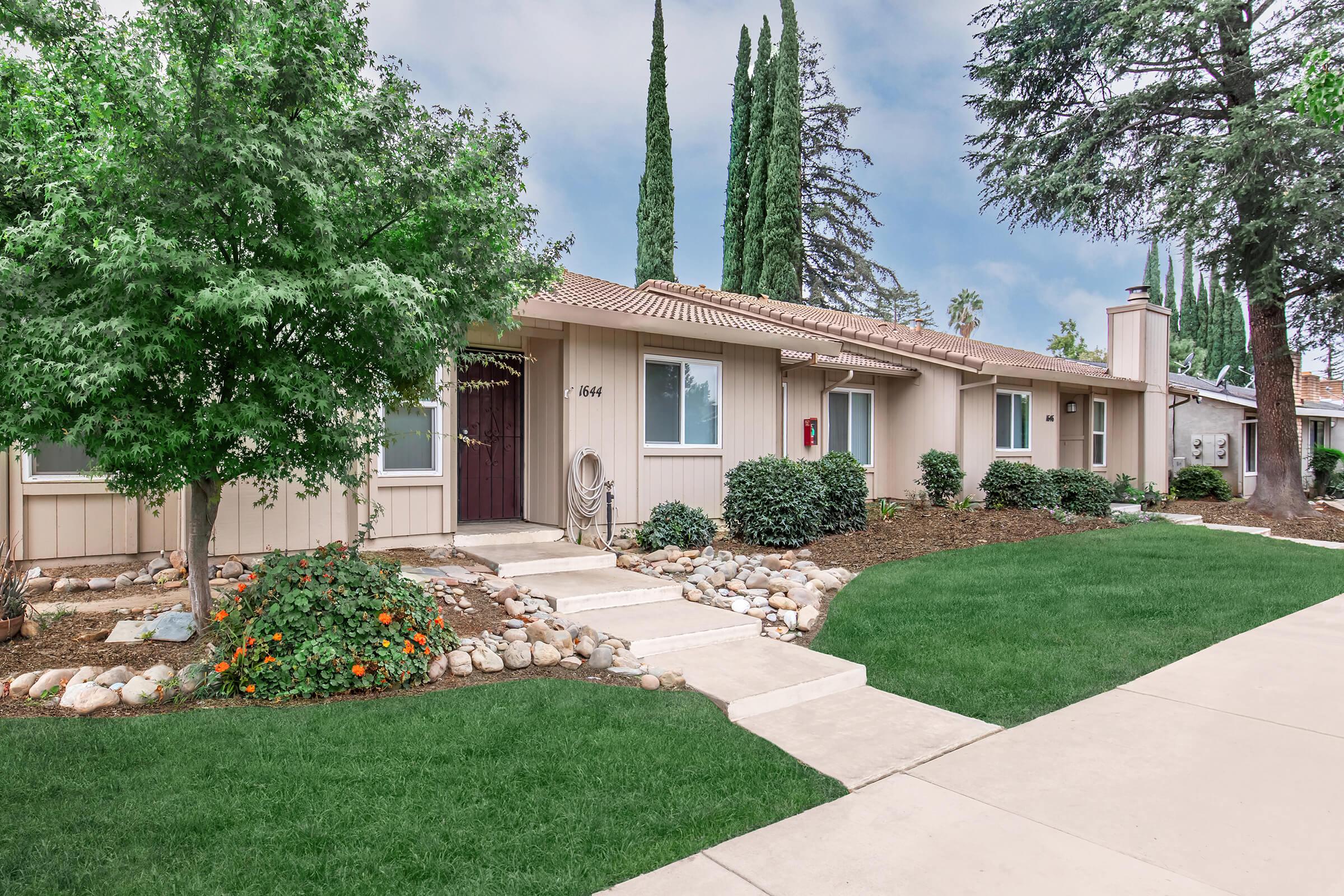 A well-maintained single-story home surrounded by lush green lawns and decorative stone landscaping. The house features a light-colored exterior, a brown door, and is framed by tall, slender trees. A concrete pathway leads to the entrance, offering an inviting atmosphere in a residential neighborhood.
