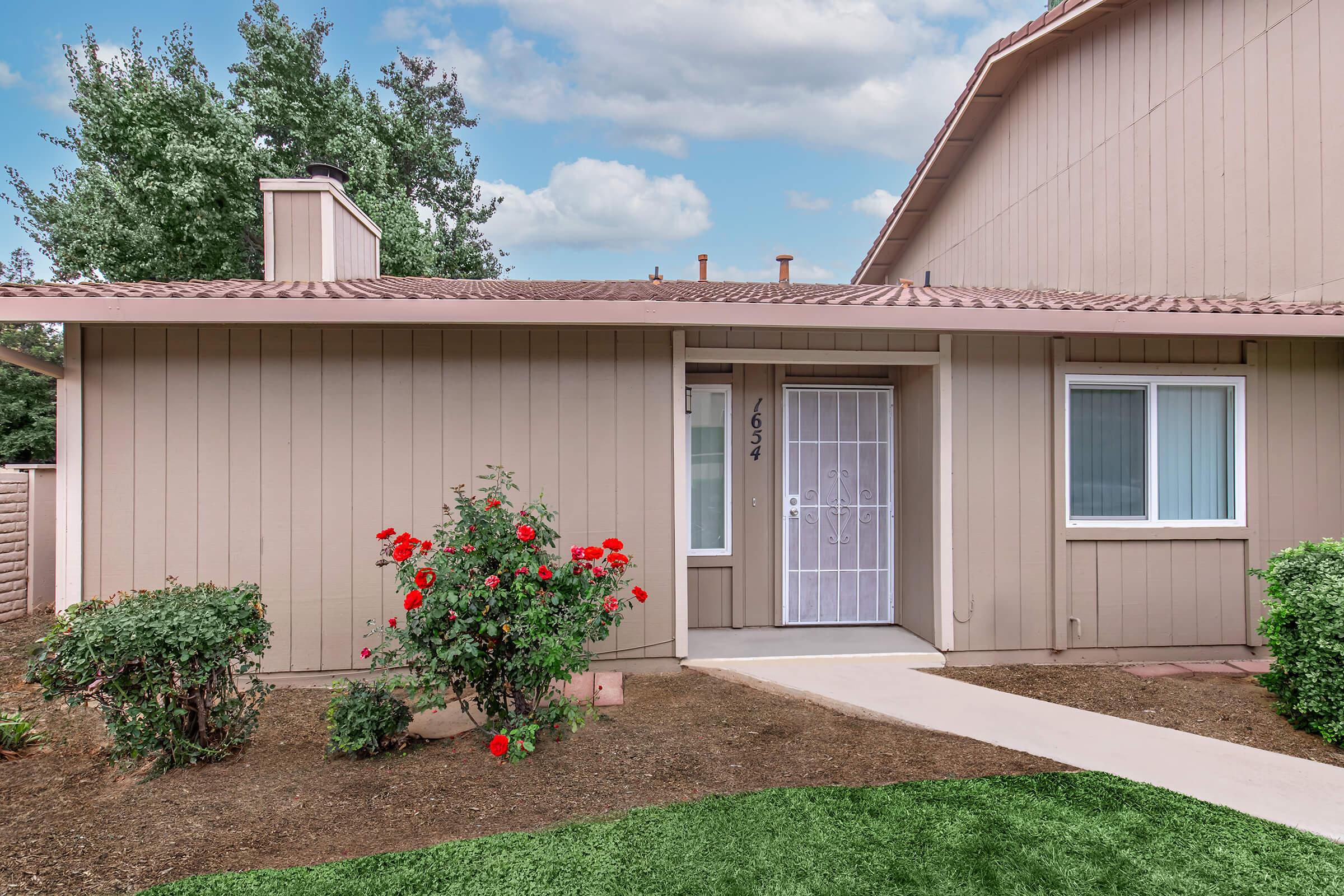 A cozy single-story home with light brown siding and a tiled roof. The front yard features vibrant red roses and small shrubs, with a clear walkway leading to the front door, which is adorned with a decorative screen. The background includes a blue sky and fluffy clouds.