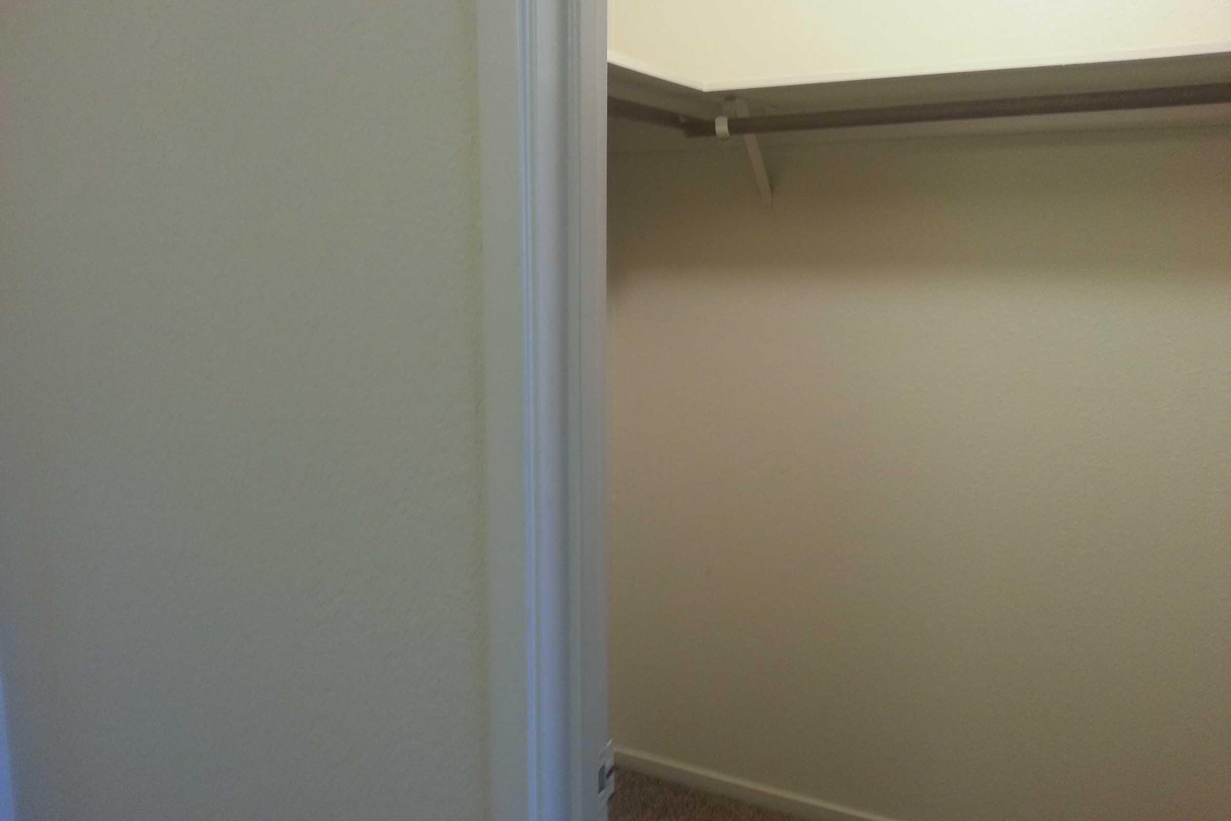 A view of a small, empty closet with beige walls. The closet is open, revealing a shelf and a hanging rod, and is located next to a white doorframe. The floor is carpeted, and the lighting is soft, giving a neutral and clean appearance.