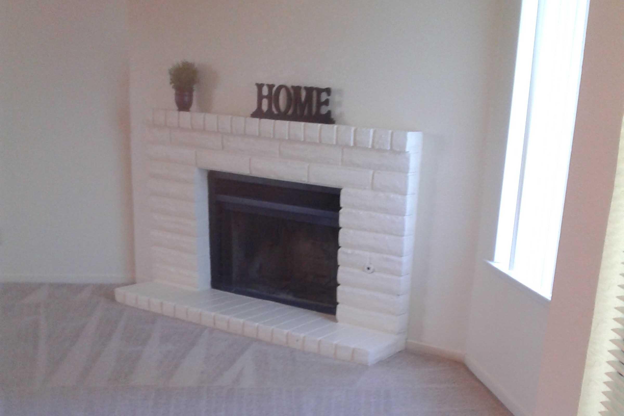 A white brick fireplace with a black frame, featuring a small plant on top and the word "HOME" displayed prominently. The area around the fireplace is carpeted, and there's natural light coming in from a window nearby.