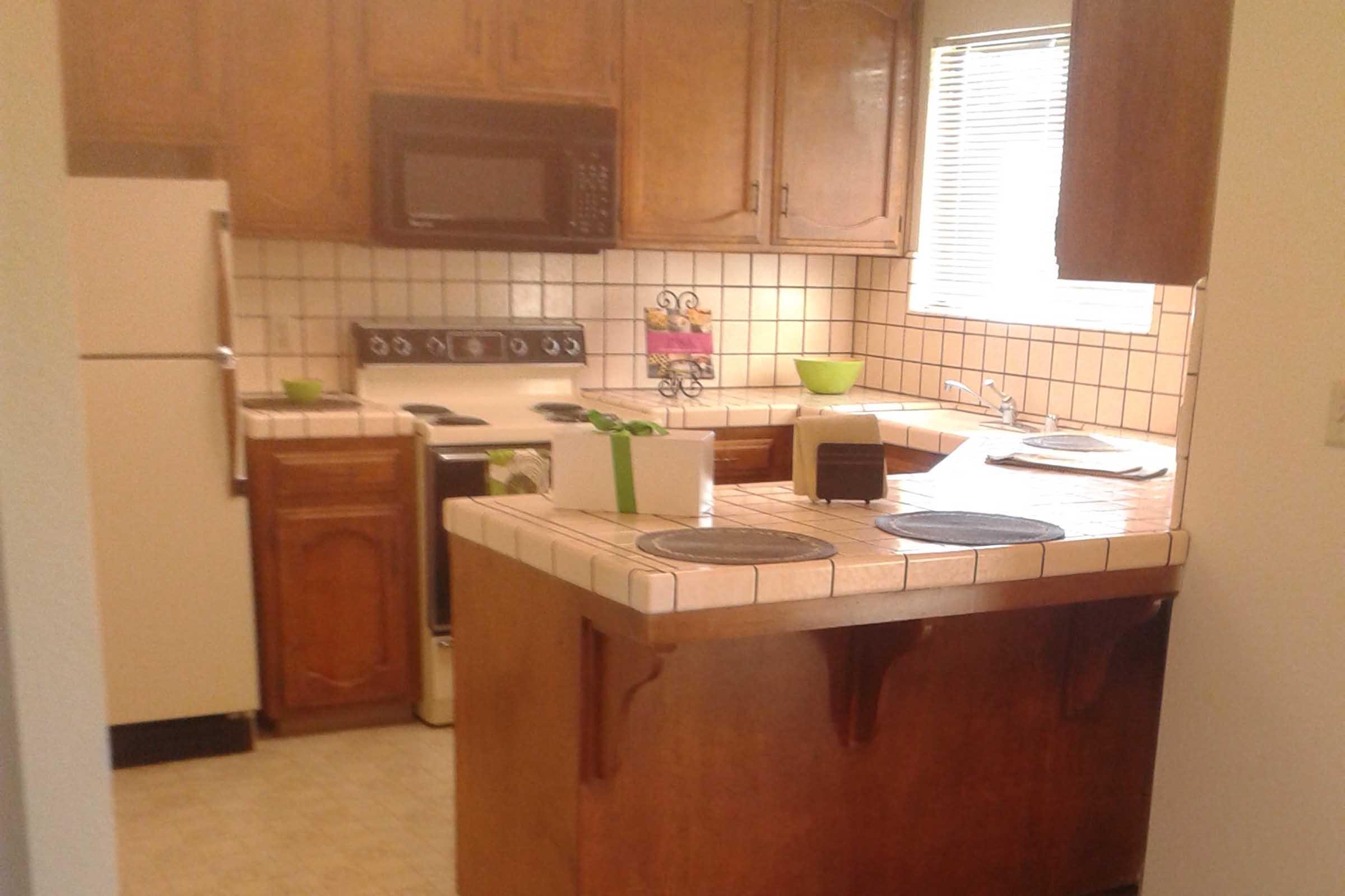 A well-lit kitchen featuring wooden cabinets, a countertop with a few appliances, including a microwave and stove. There’s a white refrigerator, a sink by the window with natural light coming in, and decorative bowls on the counter. The tile backsplash adds a classic touch to the space.
