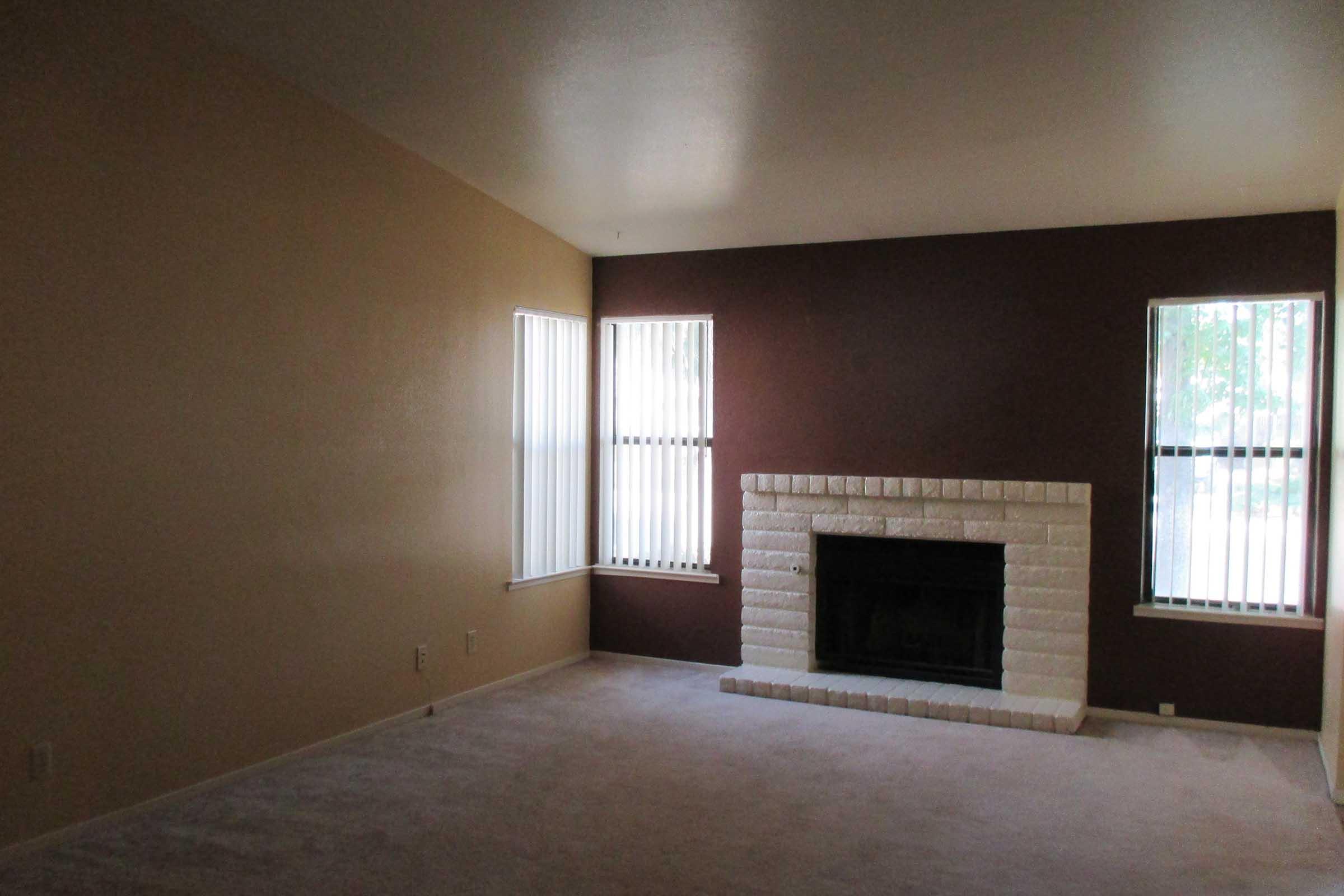 Spacious living room with a beige and brown color scheme, featuring two tall windows with vertical blinds, and a white brick fireplace as the focal point. The room has light carpet and a neutral wall color, creating a bright and airy atmosphere.