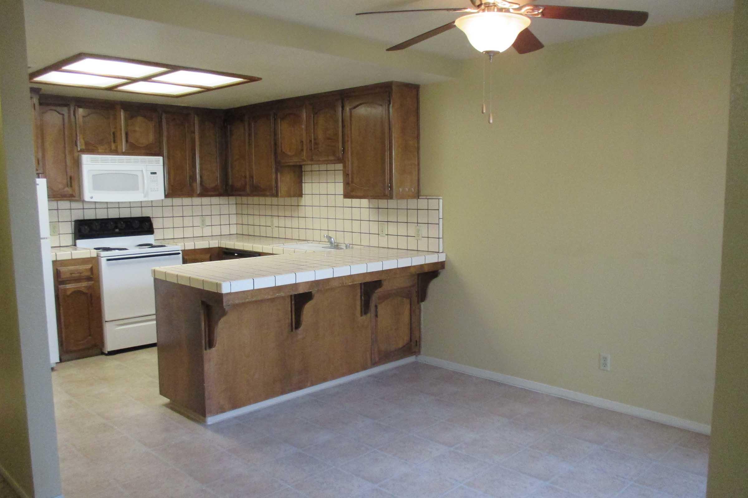 A simple kitchen featuring wooden cabinets and a tiled countertop. There is a white stove and microwave against the wall, with a ceiling fan above. The walls are painted a light yellow, and the floor is tiled in a neutral color, creating a bright and open space.