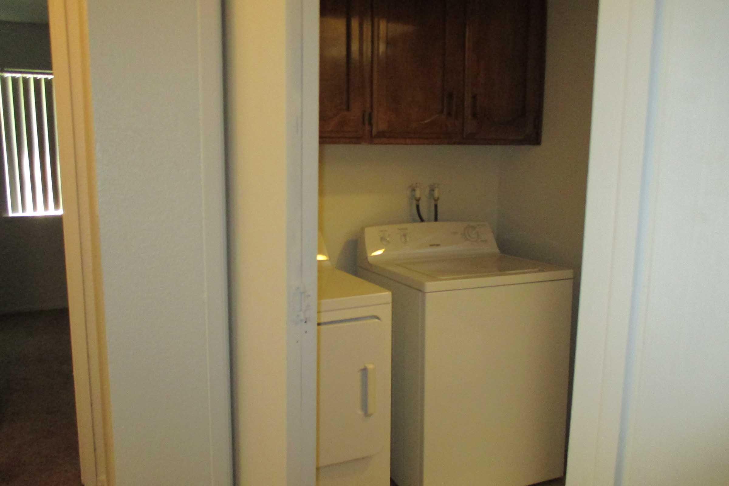 A small laundry room visible through an open doorway, featuring a washing machine and a dryer. The walls are painted in a light color, and there are wooden cabinets above the appliances. Natural light comes in from a window, casting soft shadows in the space.