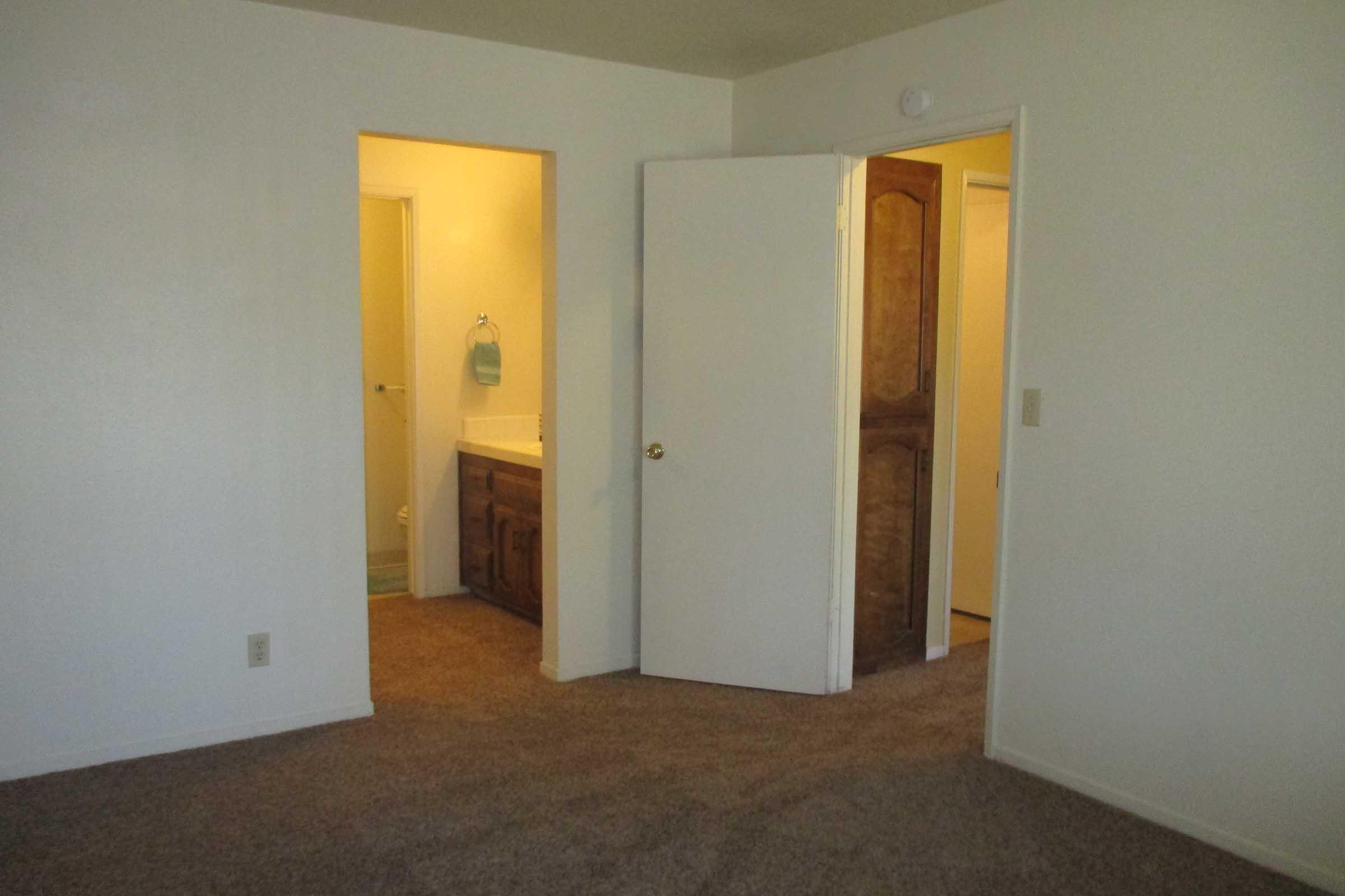 A simple interior view of a room featuring beige walls and carpet. There are two doorways: one leading to a bathroom with a sink and the other to an adjacent room. The lighting is soft, creating a warm atmosphere. No furniture is visible.