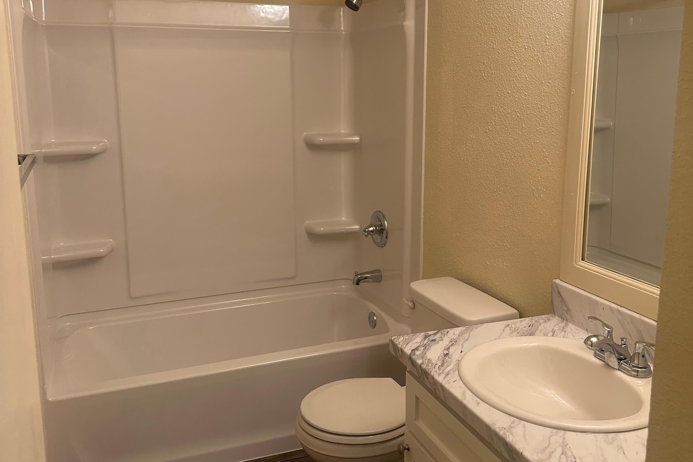 A clean and simple bathroom featuring a white bathtub with a showerhead, a toilet, and a sink with a marble countertop. The walls are painted in a soft shade, and there's a mirror above the sink, enhancing the bright, airy feel of the space. Shelves are present in the shower area.