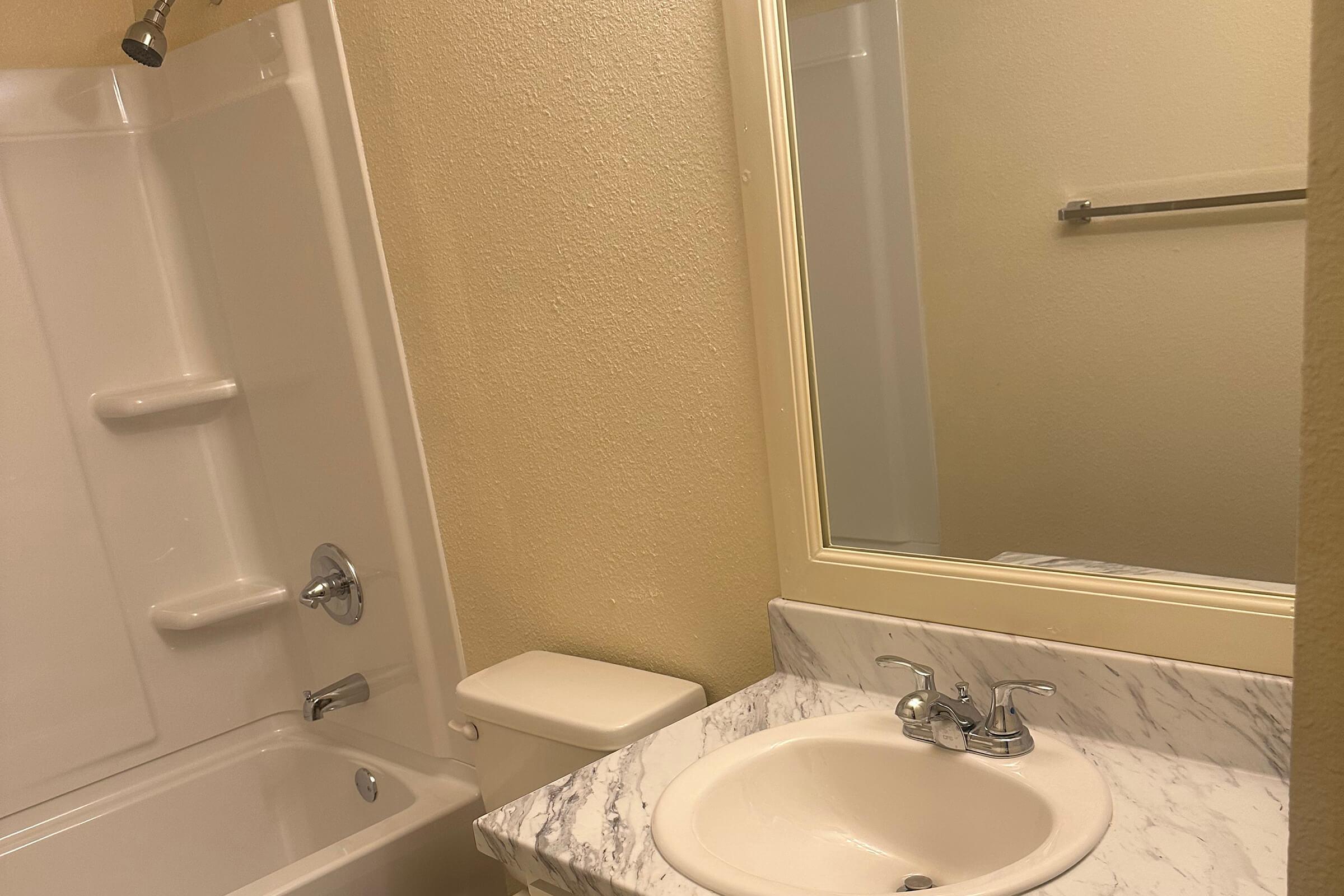 A small bathroom featuring a bathtub with a showerhead, a toilet, and a sink with a marble countertop. The walls are painted beige, and there is a mirror above the sink. A towel bar is mounted on the wall near the sink.