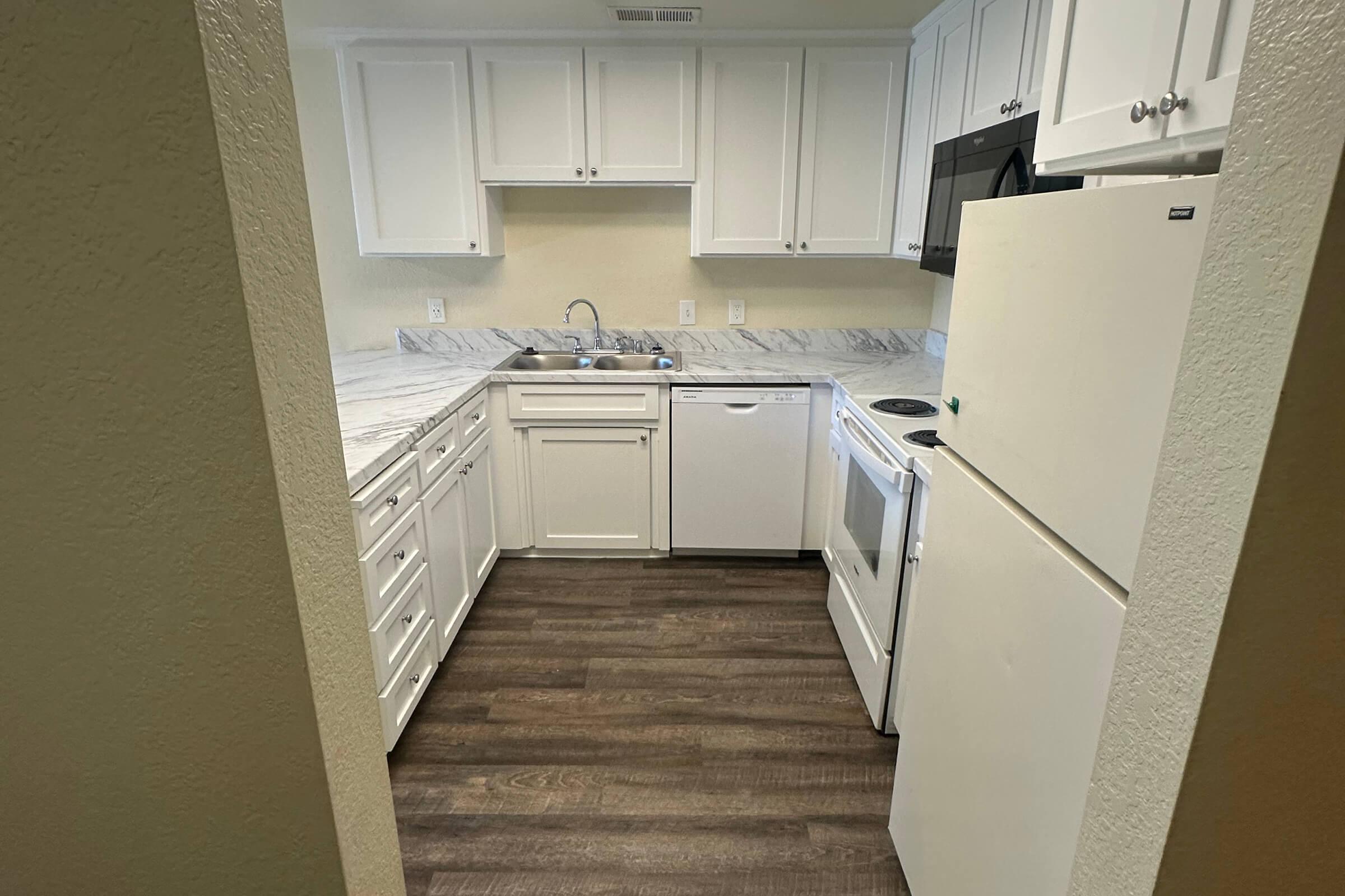 A clean, modern kitchen featuring white cabinetry, a marble countertop, a sink, a dishwasher, and a stove. The floor has a wood-like appearance, and the kitchen is well-lit with light-colored walls. An entranceway leads into the kitchen from a hallway.