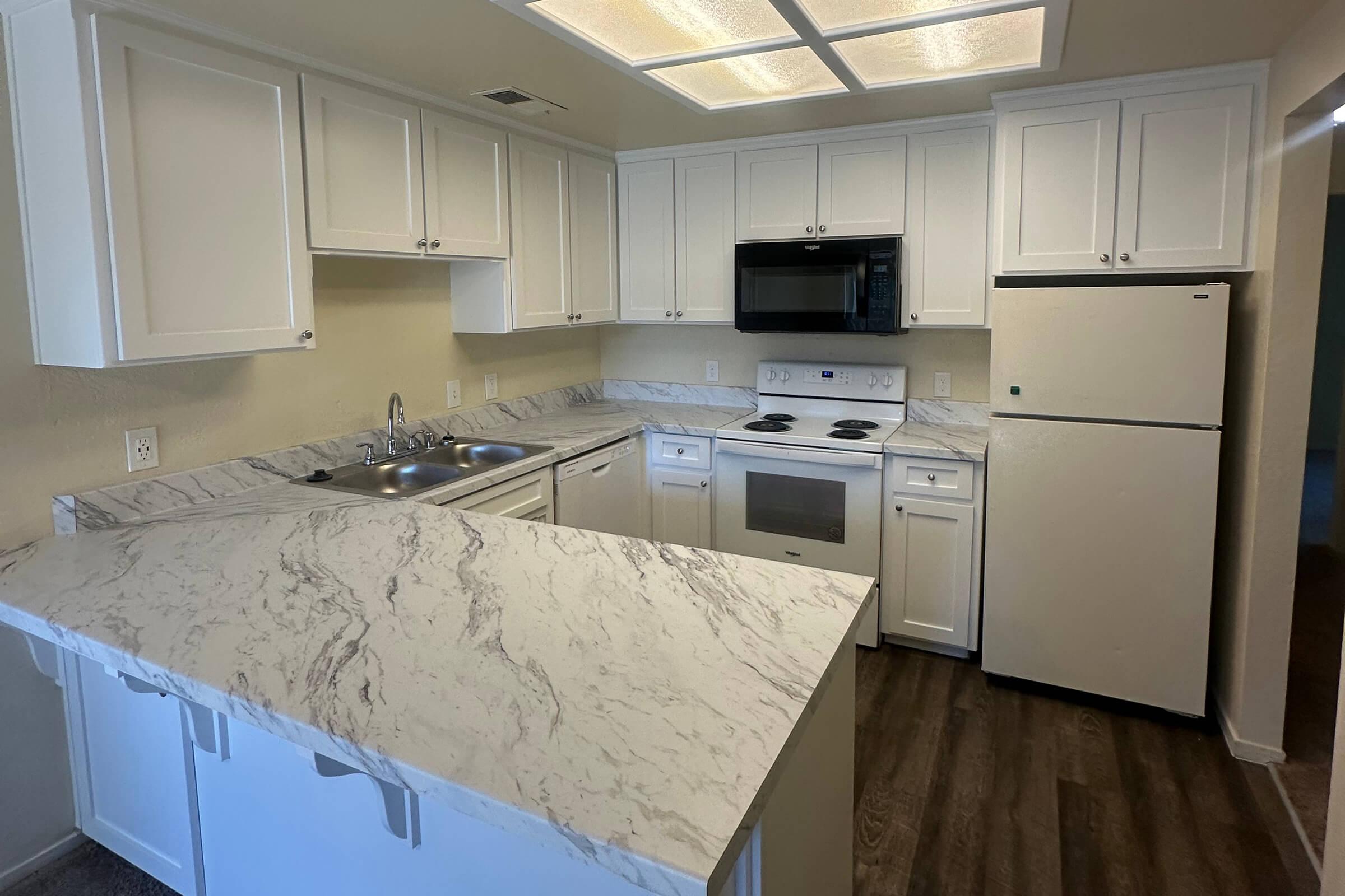 A modern kitchen featuring white cabinetry, a marble countertop, a double sink, and a black microwave above the stove. The kitchen has stainless steel appliances, including a refrigerator and an oven, with recessed lighting illuminating the space. The flooring is wood-like laminate.