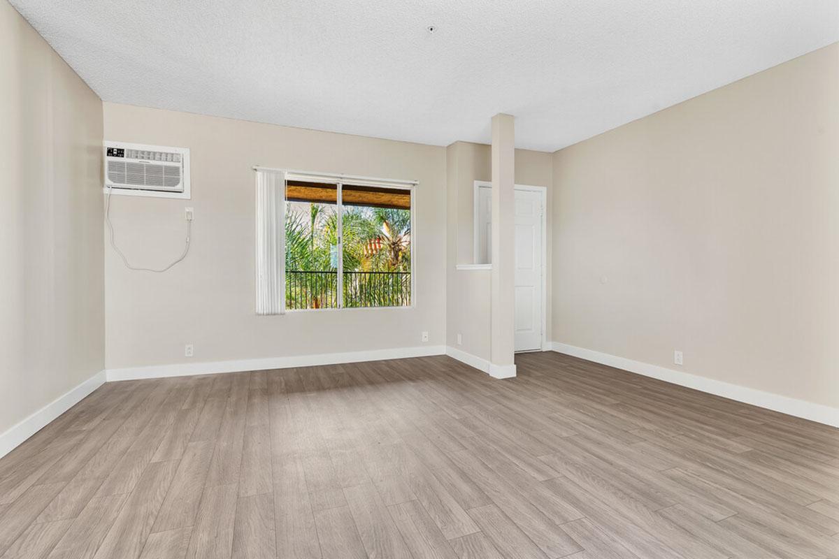 A spacious, empty living room featuring light-colored walls, a wooden floor, and a large window with a view of greenery. An air conditioning unit is installed on the wall. The room has a simple and modern design, providing ample natural light and a clean, uncluttered atmosphere.