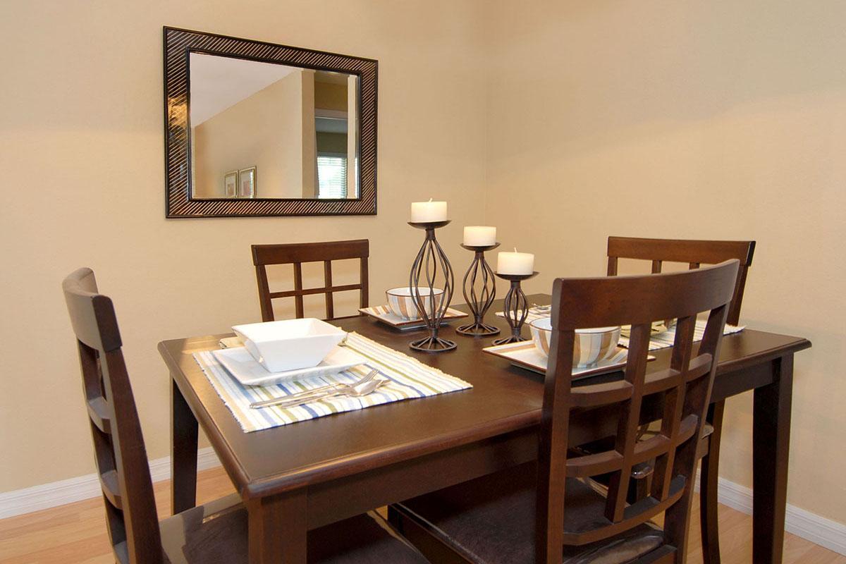 A well-set dining table with dark wooden chairs, featuring white plates, striped napkins, and decorative candles in elegant holders. A mirror hangs on the wall, reflecting the room, which has neutral-colored walls and wooden flooring.