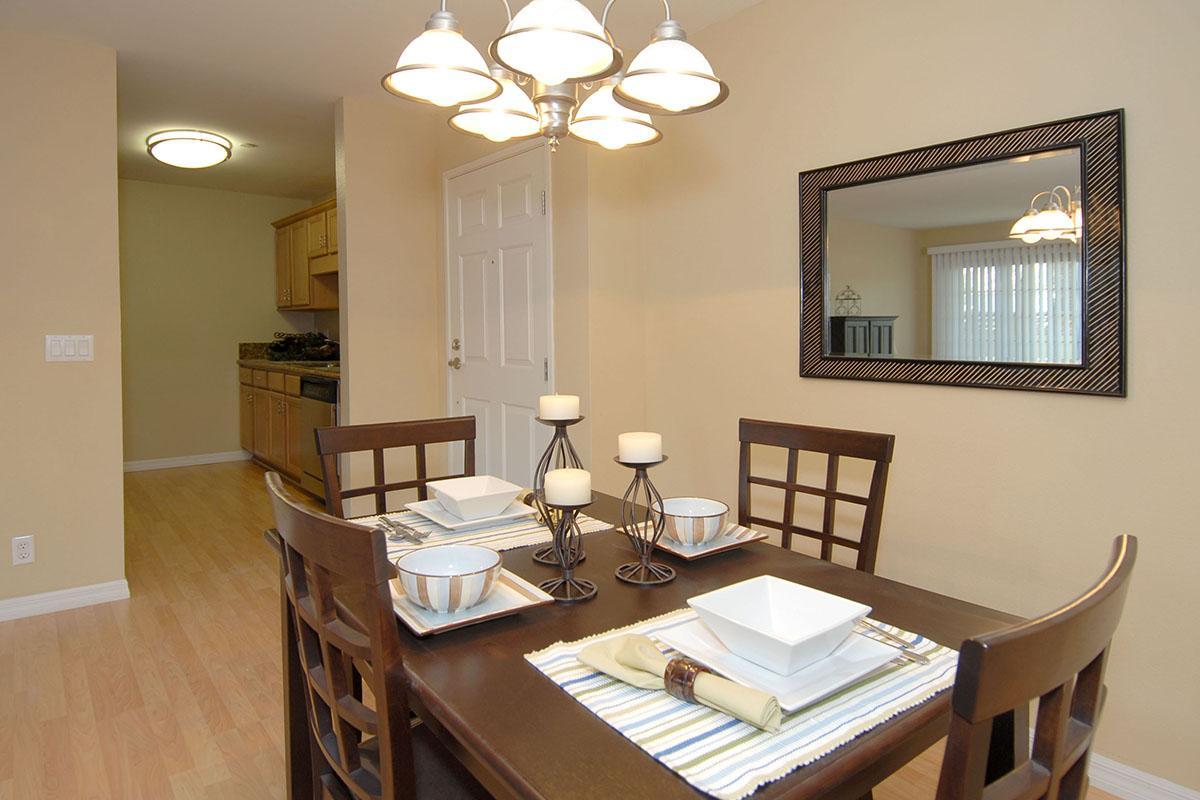 A cozy dining area featuring a dark wooden table set with white plates and candles, illuminated by a pendant light. There are four chairs around the table, with a light-colored wall and a large mirror reflecting the space. In the background, a kitchen area is visible, adding to the inviting atmosphere.