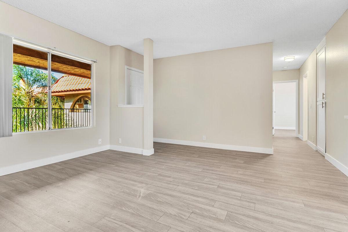 Spacious, unfurnished interior of a modern room with light-colored walls and wood-like flooring. Large window providing natural light with a view of greenery outside. Adjacent empty area that leads to another room, highlighting a bright and airy atmosphere.