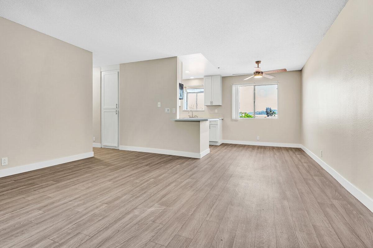 A spacious, modern living area with light-colored walls and wood-like flooring. The open layout includes a kitchen area with white cabinetry, a window allowing natural light, and a ceiling fixture. The room feels bright and airy, complemented by a neutral color palette.