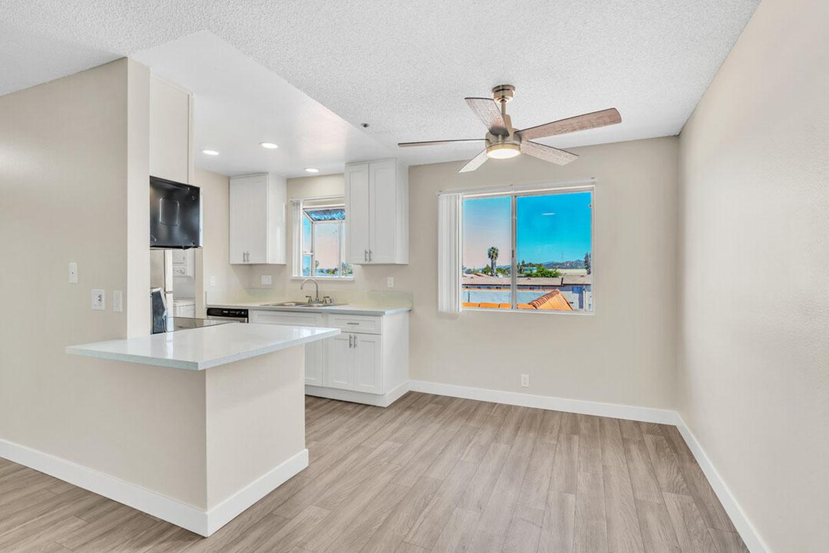Bright and spacious modern kitchen with white cabinetry and a light countertop, featuring a ceiling fan above. A window allows natural light to enter, revealing a view of the outdoors. The floor has light wood-like tiles, creating a warm atmosphere in a freshly painted neutral-toned room.