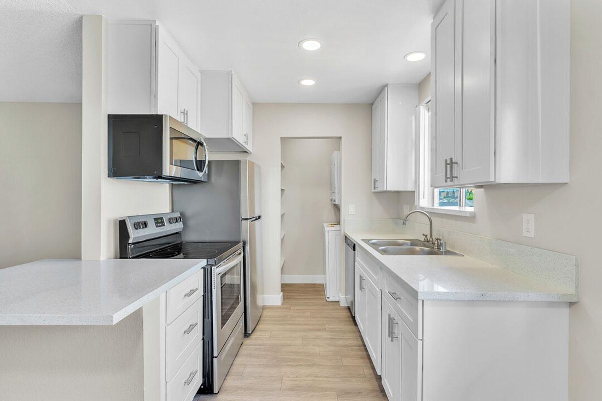 Modern kitchen featuring stainless steel appliances, including a microwave and oven, quartz countertops, and white cabinetry. There is a sink under a window, light-colored wood flooring, and an adjacent space with a door, possibly for a pantry or laundry. Brightly lit with recessed lighting.