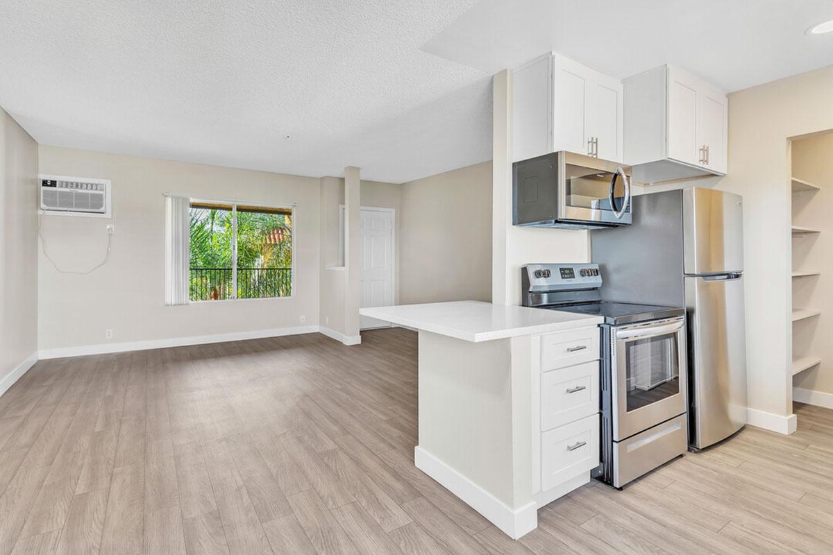 Bright, modern kitchen and living area featuring stainless steel appliances, white cabinetry, and a large window with greenery outside. The space has light-colored walls and a wood-style floor, creating an inviting atmosphere. Air conditioning unit visible in the corner.