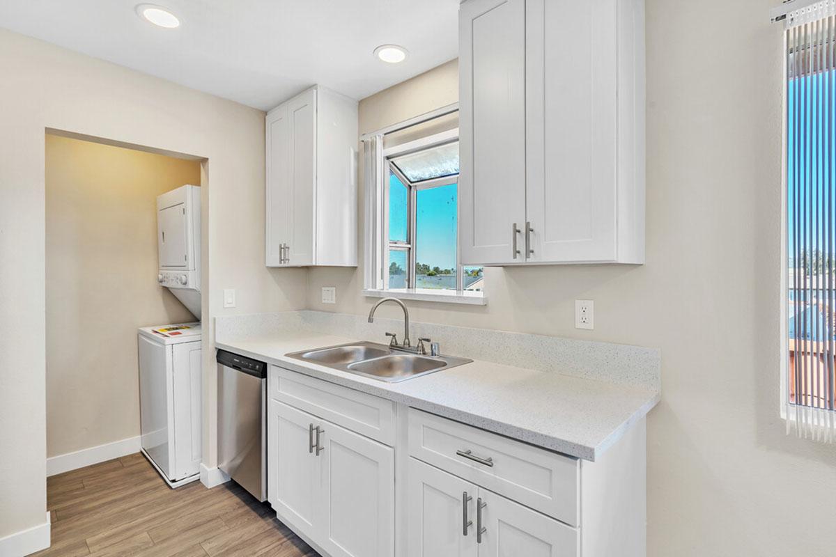 A modern kitchen featuring white cabinetry, a double sink, and a dishwasher. There's a laundry area visible in the background with a washer and dryer. Natural light enters through a window above the sink, and the flooring is a light wood grain.