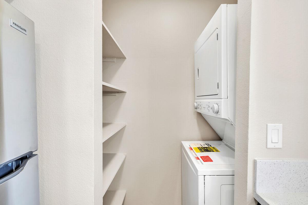 A small laundry area featuring a stacked washer and dryer unit next to open shelves against a beige wall. There is a light switch on the right side, and a stainless steel refrigerator is partially visible on the left. The overall space appears neat and organized.