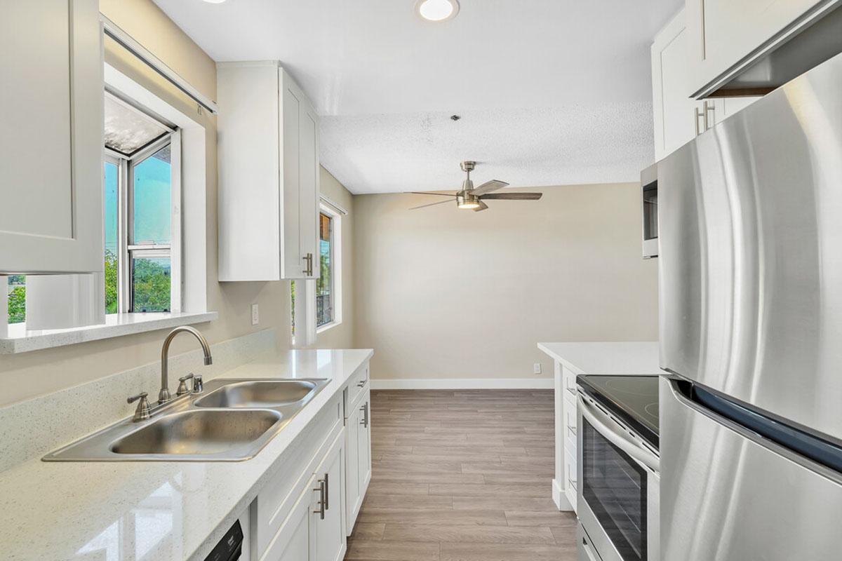 Modern kitchen with a clean design featuring white cabinetry, a stainless steel stove and refrigerator, and a double sink. The room has large windows allowing natural light and a ceiling fan for ventilation. Light-colored walls and wooden flooring enhance the spacious feel.