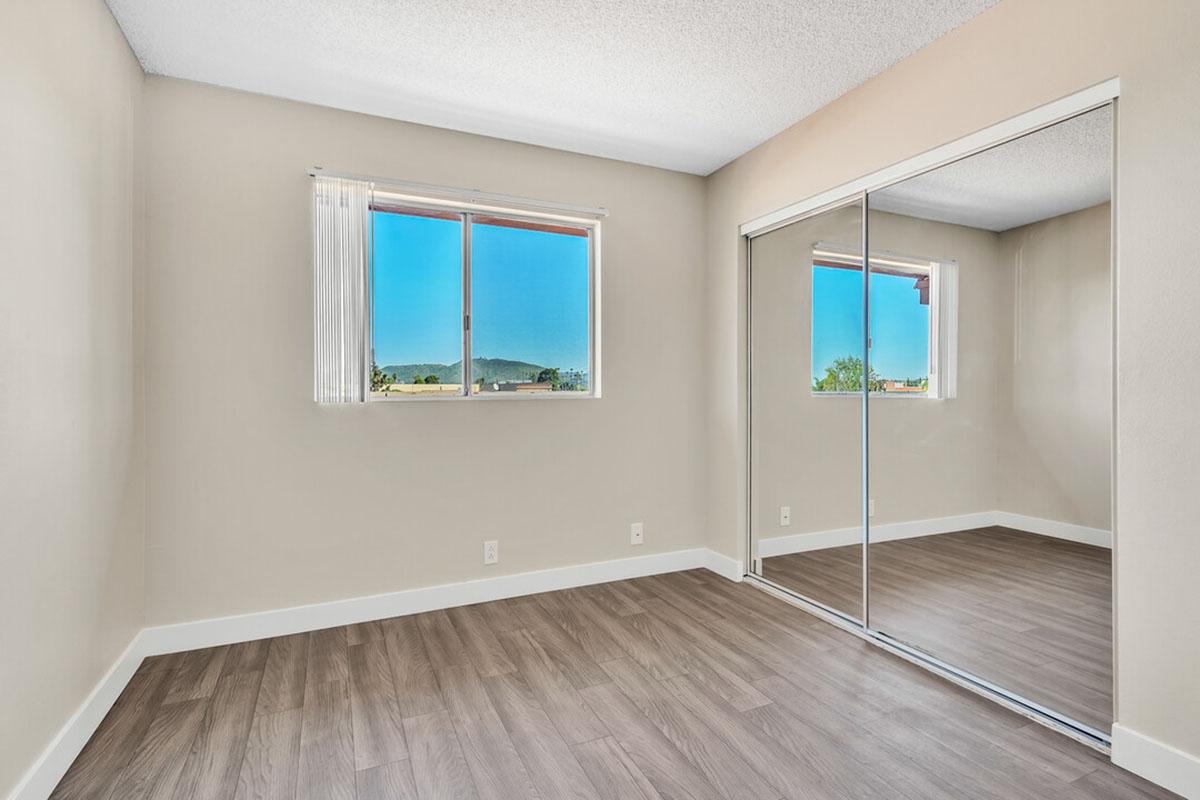 A minimalist bedroom featuring light-colored walls and a large window with a view of mountains. The room has a wooden floor and a mirrored closet that reflects the space, enhancing the natural light. The overall design is simple and modern, creating a calm atmosphere.
