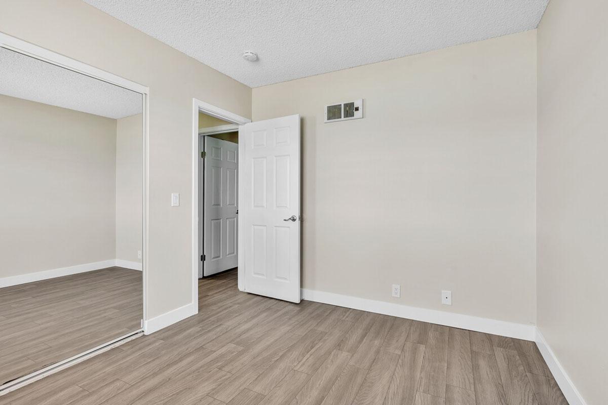 A clean, empty room featuring light-colored walls, a mirror closet door, and a standard door partially open to another room. The flooring is light wood, and the ceiling has a textured finish. Natural light brightens the space, creating a minimalist atmosphere.