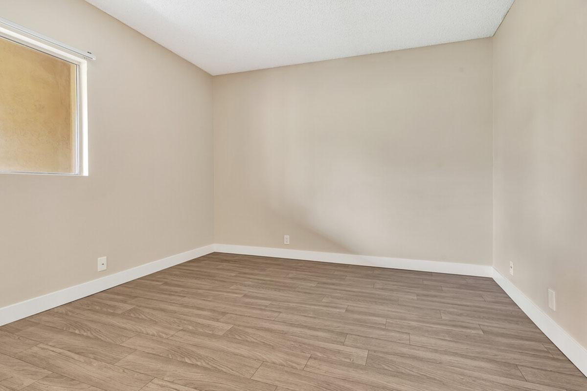 A vacant room with light brown walls and a wood-like floor. There is a single window on the left side allowing natural light to enter, and the room appears spacious and empty, ready for furnishing or decoration.